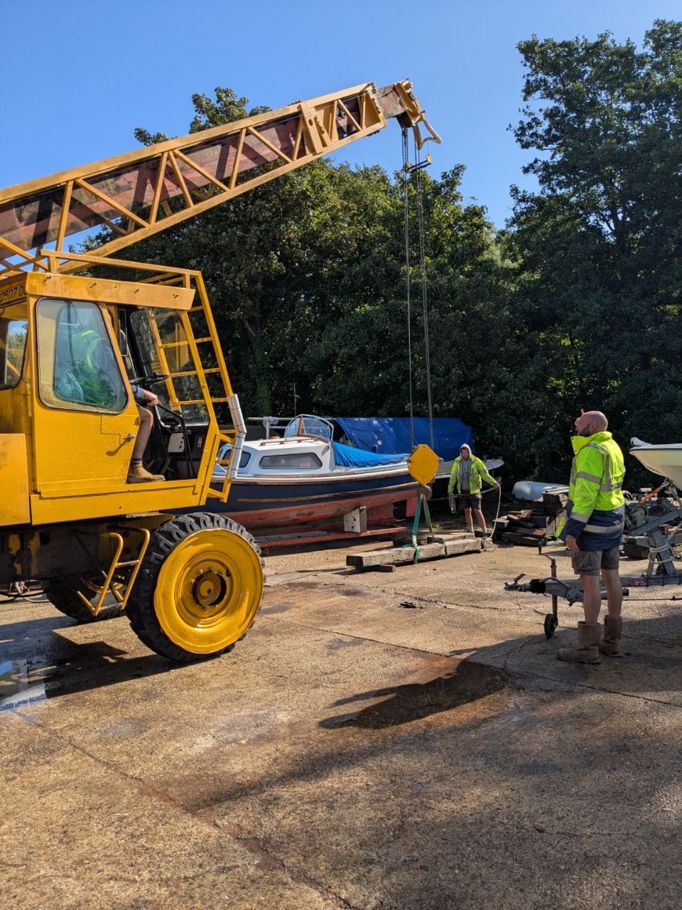 Yellow crane lifting a boat. Workers in vests stand nearby. Sunny day, outdoors.