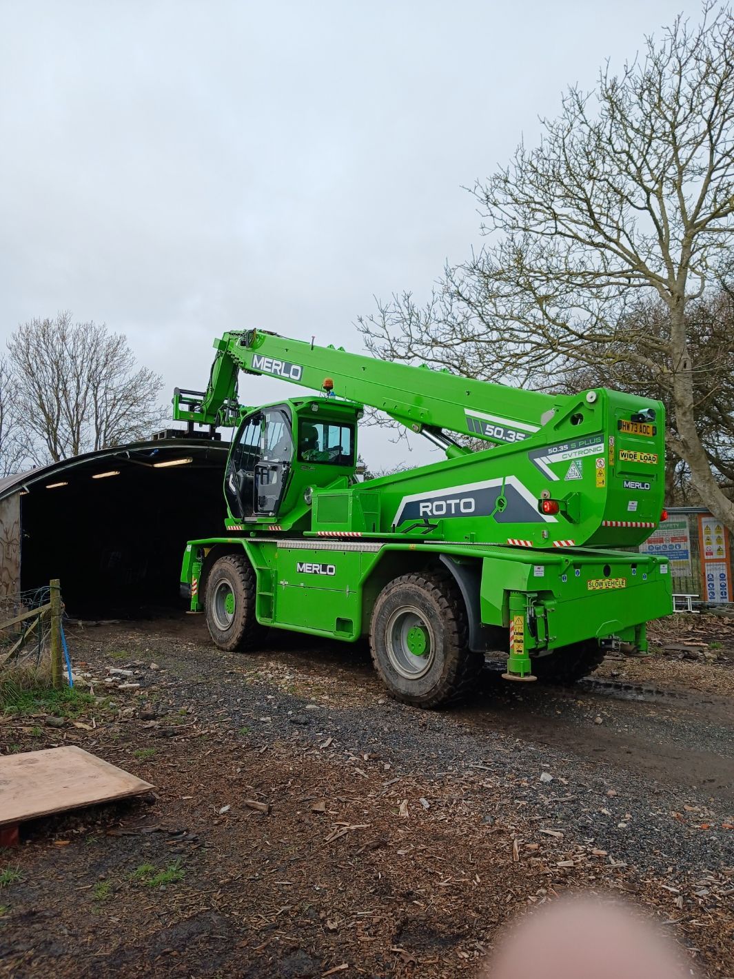 A bright green Merlo Roto telehandler parked outdoors near a dark shed on a cloudy day.