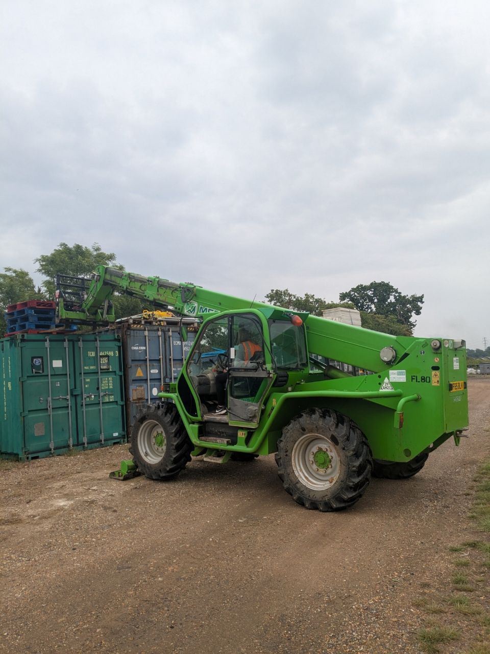 A green forklift is parked in a dirt field next to a green container.