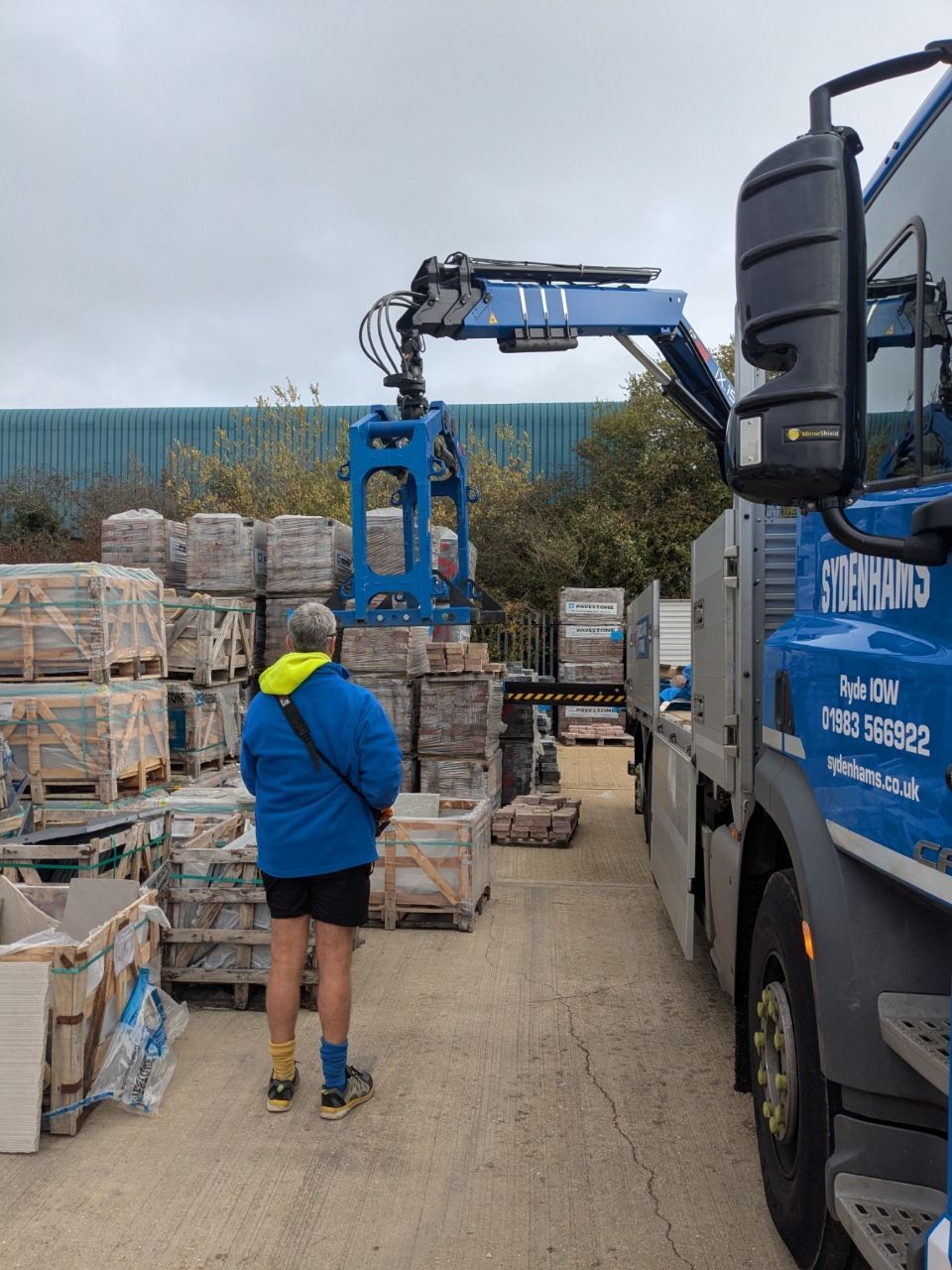 A man watches a truck-mounted crane lift crates of pavers at a construction supply yard.