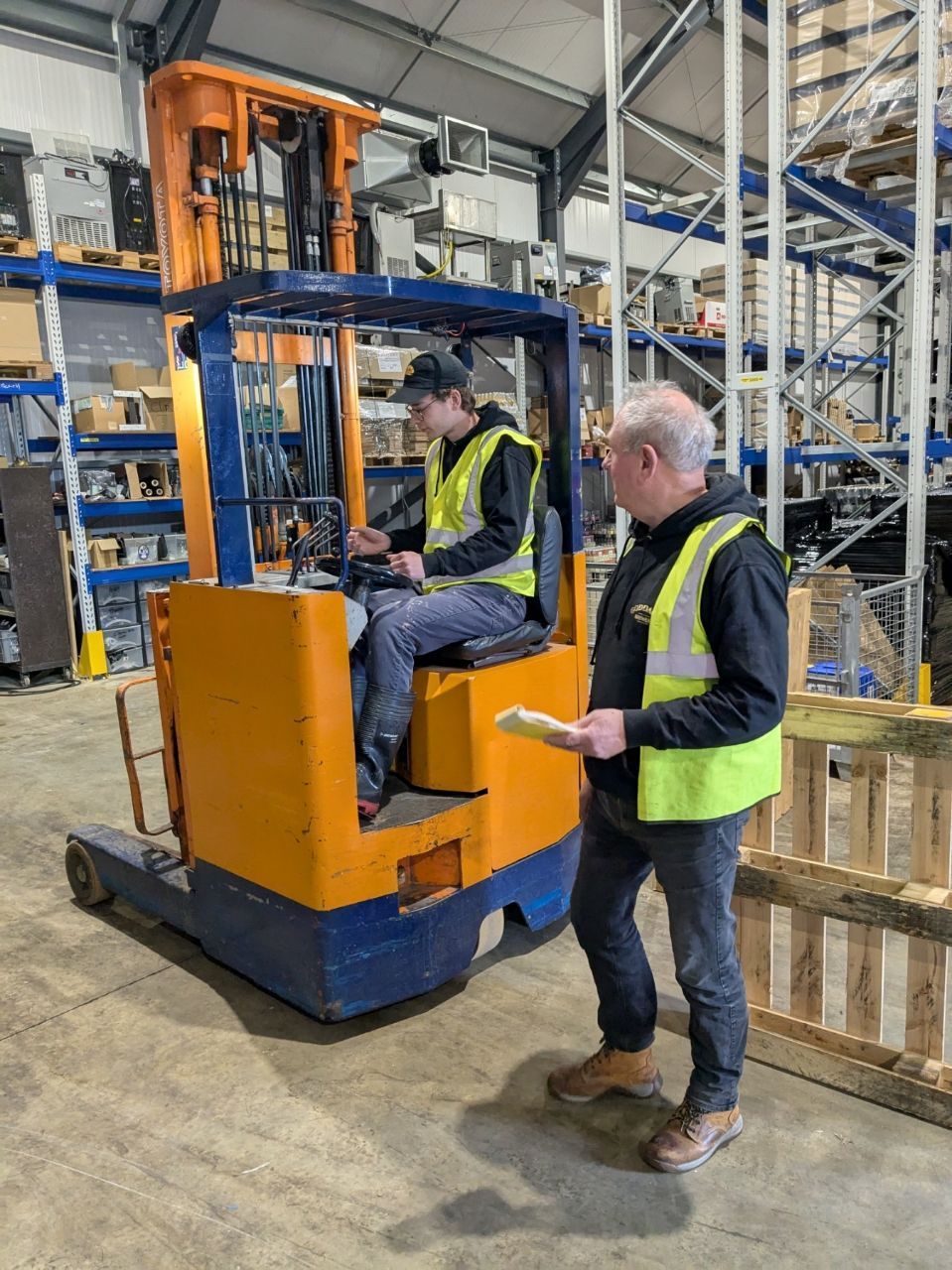 Man in yellow vest instructs another in a forklift in a warehouse; both wear vests.