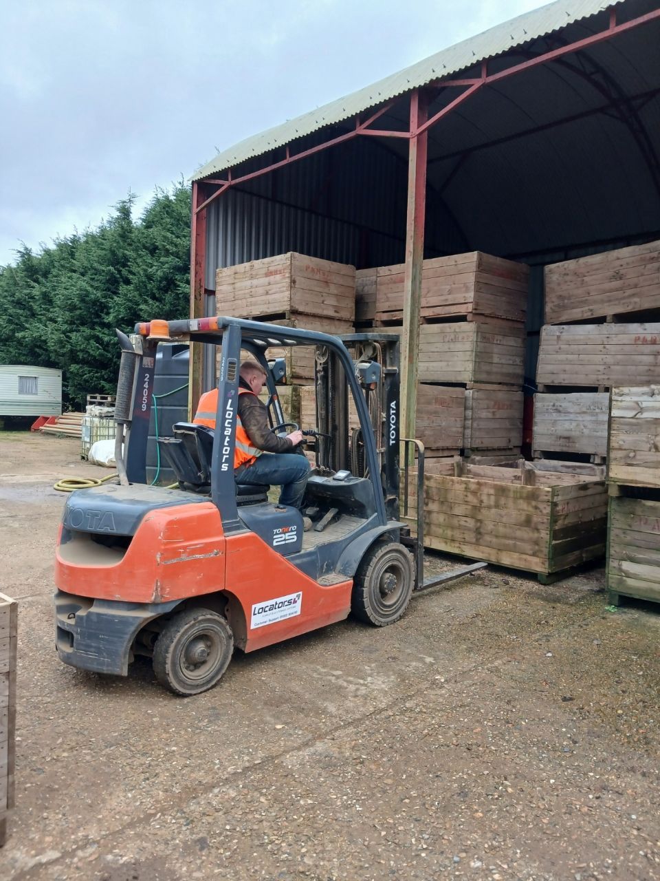 Forklift operator moving wooden pallets in a warehouse. Orange forklift, stacks of wood, cloudy sky.