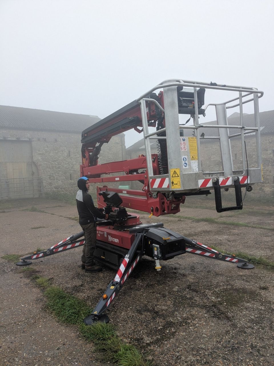 A person operating a red boom lift outdoors. It is a foggy day, near a stone building.