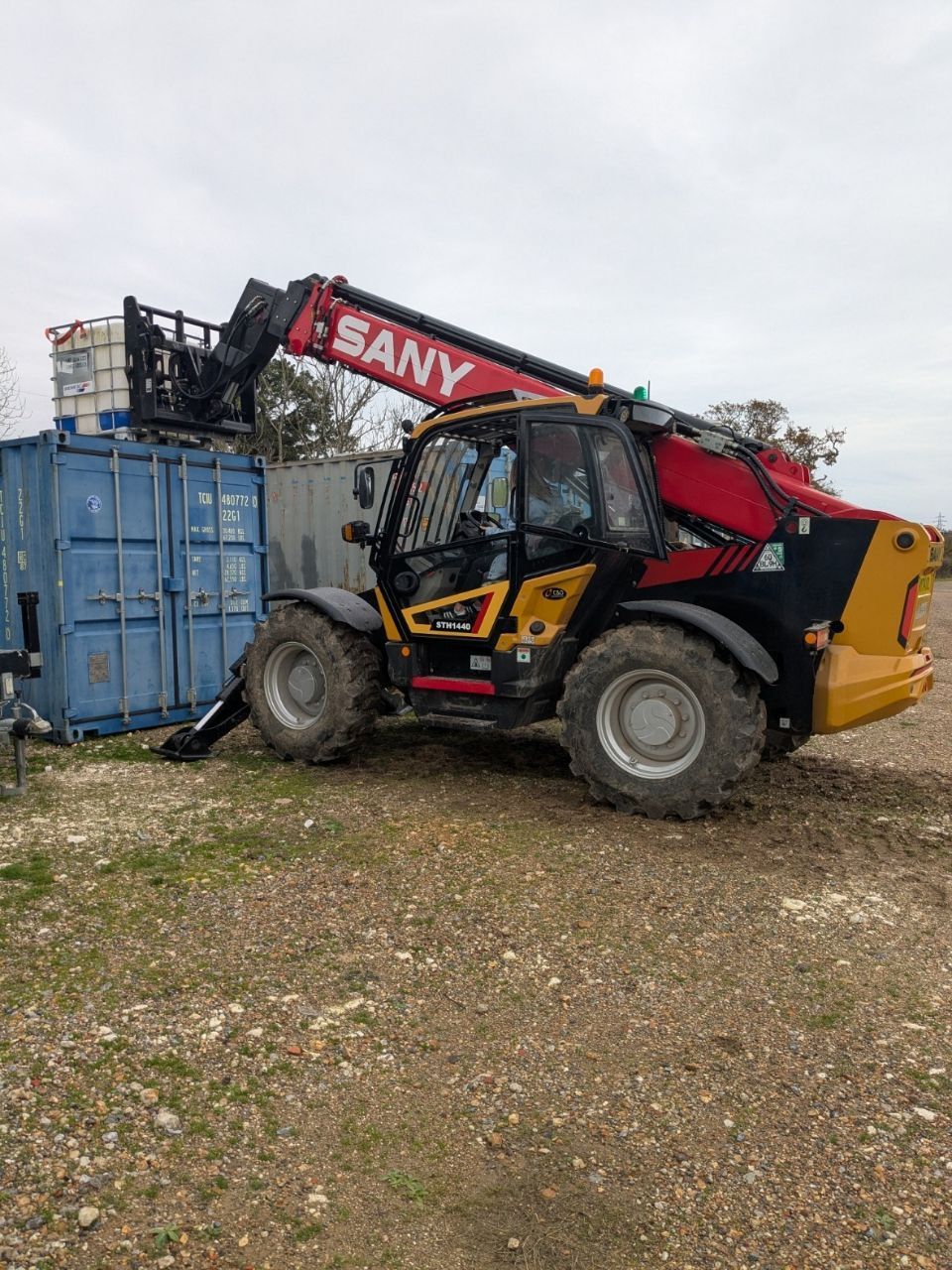 A red and yellow SANY telehandler lifting a blue shipping container in an outdoor setting.