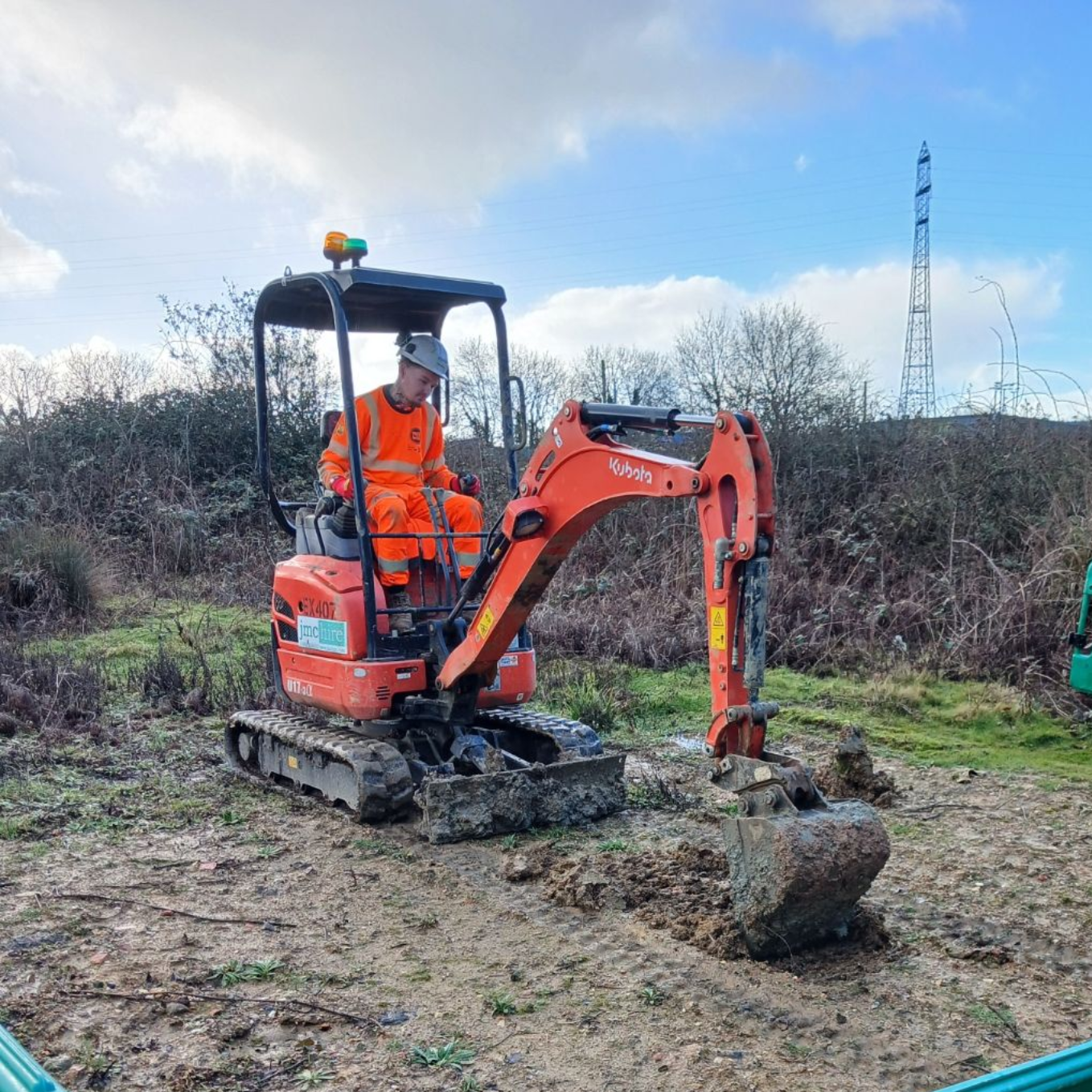 A worker in orange safety gear operates a small excavator outdoors, digging in the mud; green barriers in the foreground.