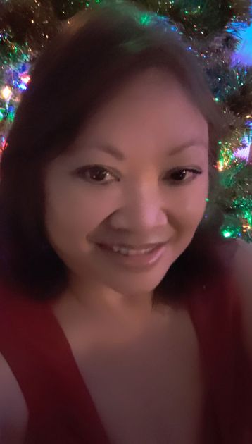 Woman smiling in front of a Christmas tree with colorful lights, wearing a red top.
