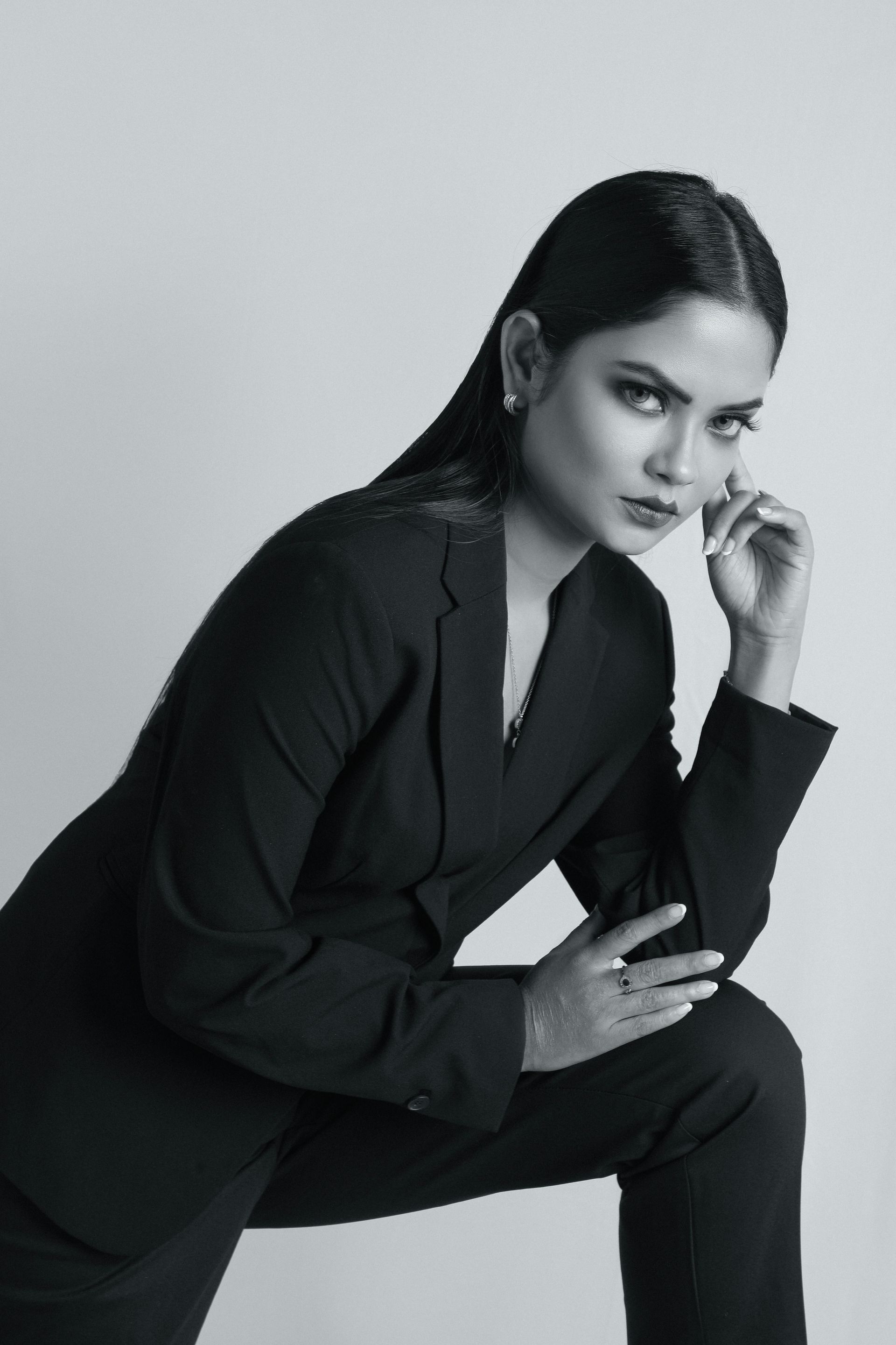 Woman in a black suit, leaning with one knee up, looking at the camera. Black and white photo.