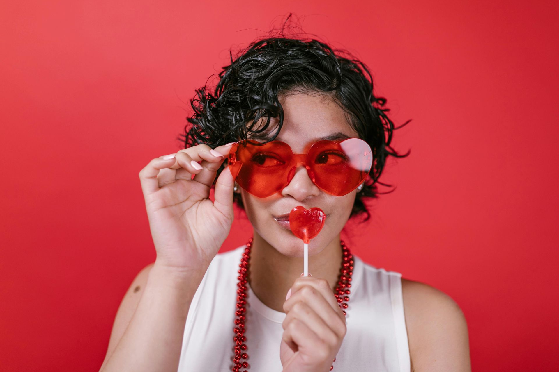 Woman with heart-shaped glasses and lollipop, holding up glasses, red background.