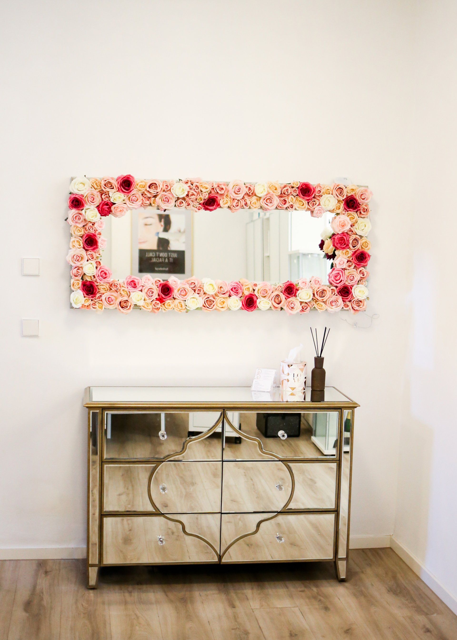 Über einer Kommode in einem Zimmer hängt ein Spiegel mit Blumen darauf.