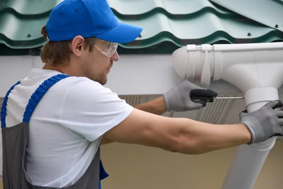 Worker in blue cap and gloves adjusting a white gutter pipe on a house exterior.