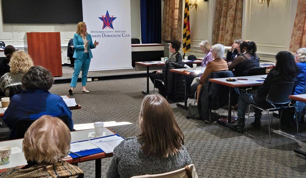 Woman in teal suit presents to a seated audience in a conference room. Banner with logo behind her.