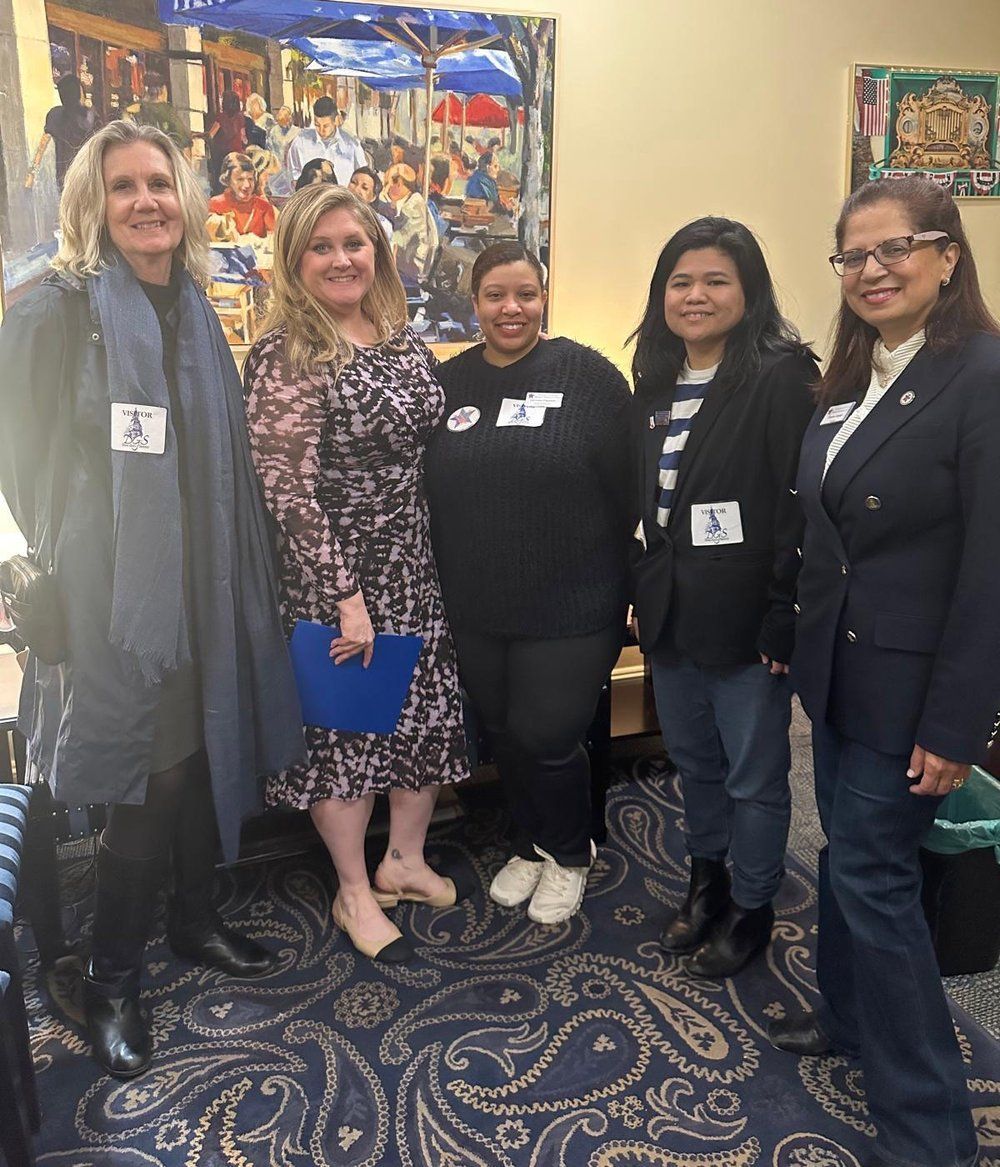 Five women pose together indoors. One wears a long gray coat, another a patterned dress, a third a black shirt, and two wear jackets. A colorful mural is behind them.