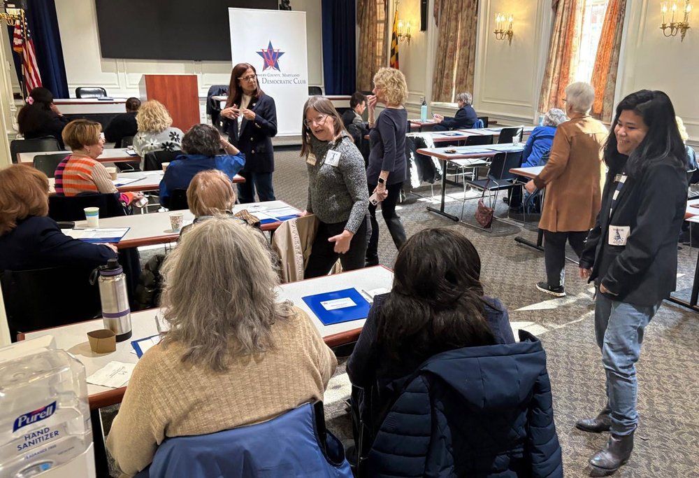 People at a conference with speakers. Attendees sit at tables; some are talking or standing.