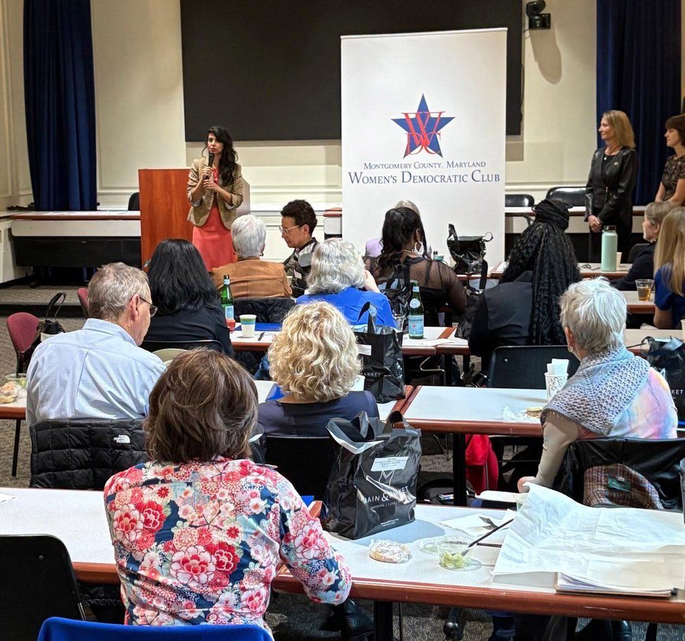 A woman speaks at a podium to a seated audience at a Women's Democratic Club event.