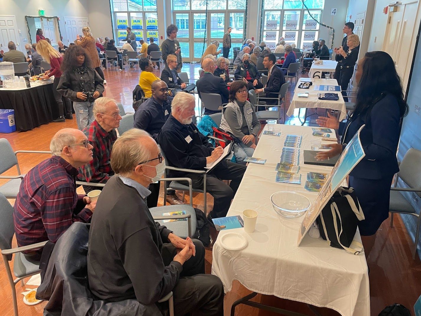 Woman giving a presentation at a community event, with attendees seated at tables in a bright hall.