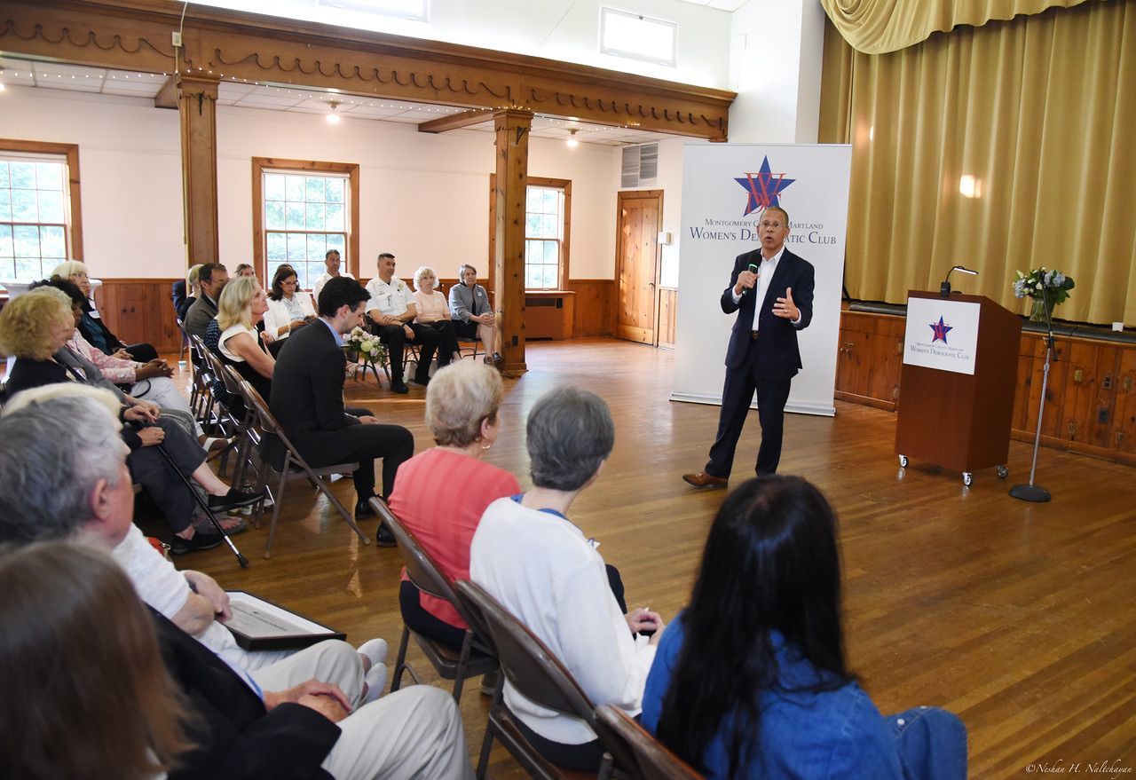 Man speaking at podium, addressing a seated audience in a wood-floored hall. Logo visible.