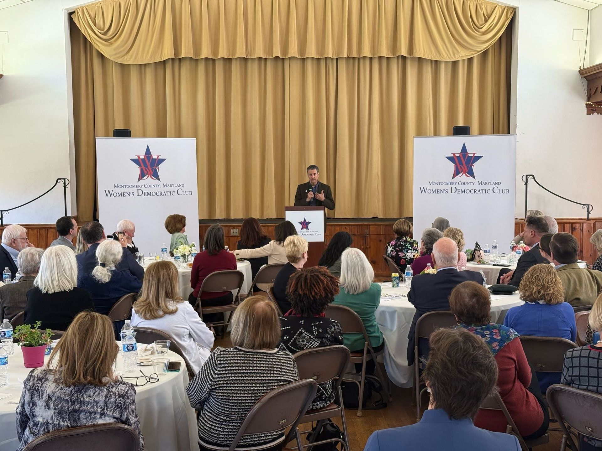Man speaking at podium for a group at tables in a decorated hall; two signs behind him.