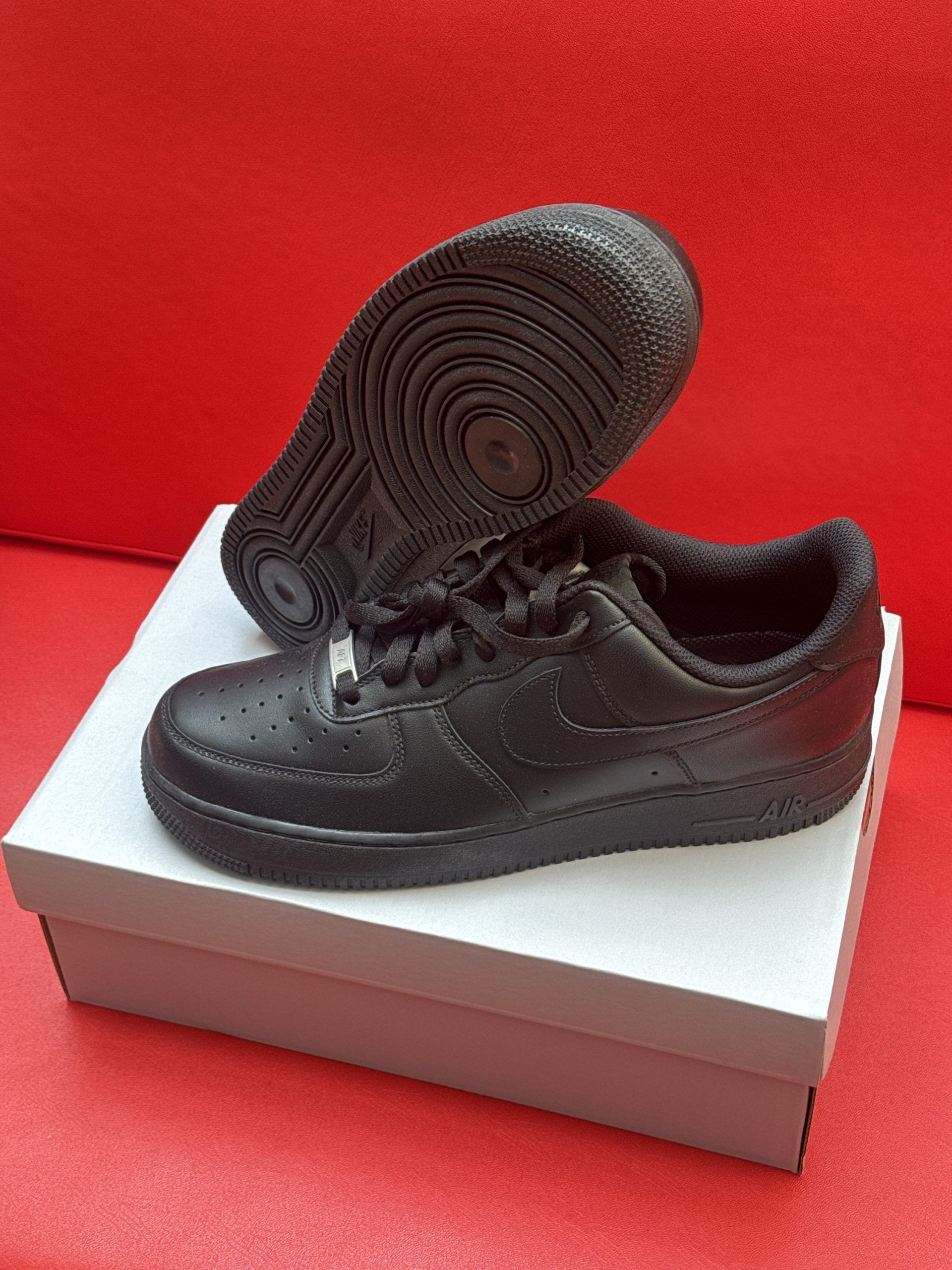 Black Nike Air Force 1 sneakers on a white box, set against a red background. The sole is visible.