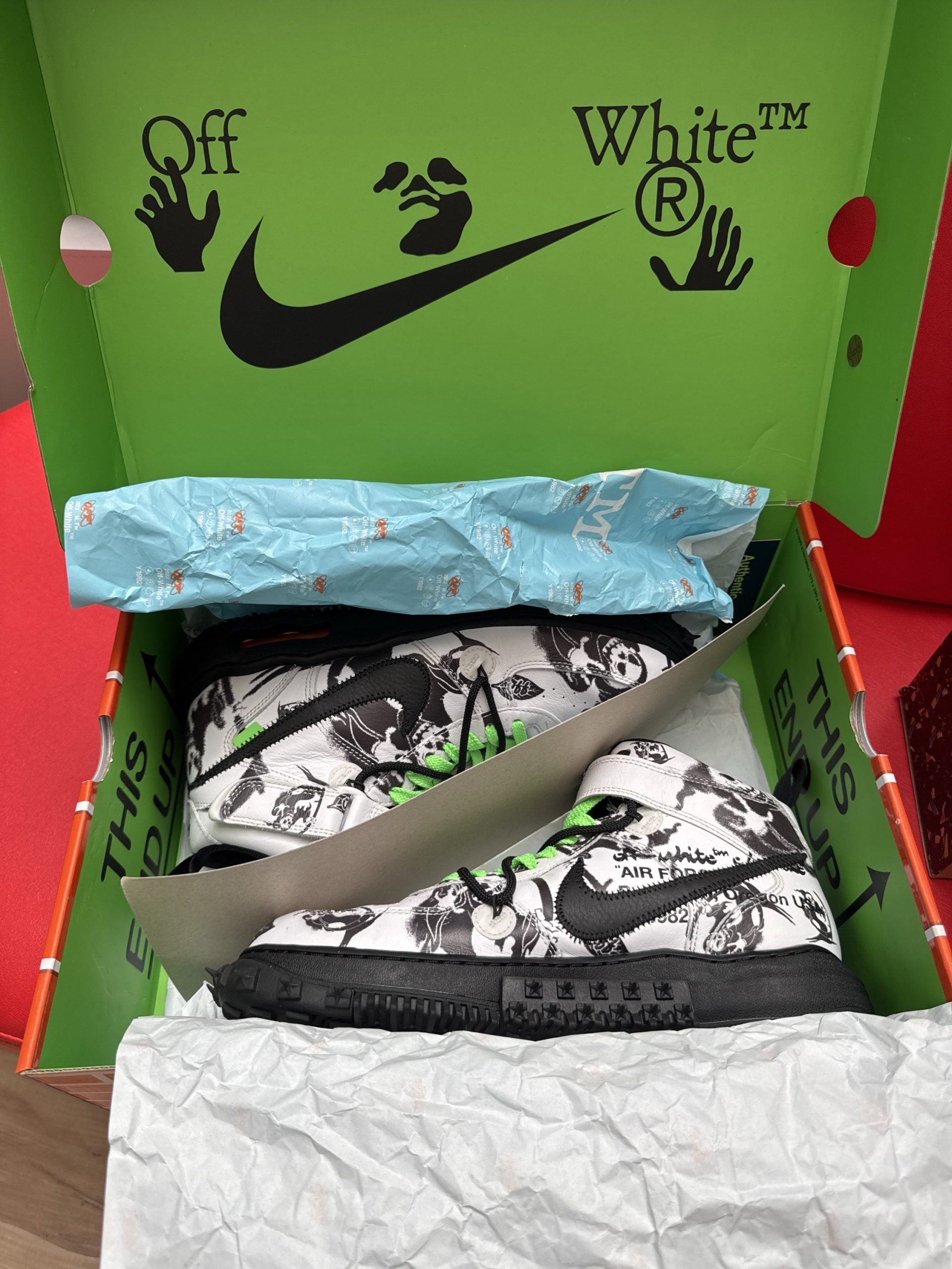 White and black patterned Nike sneaker with green laces, on a brick-patterned box, with black sole.