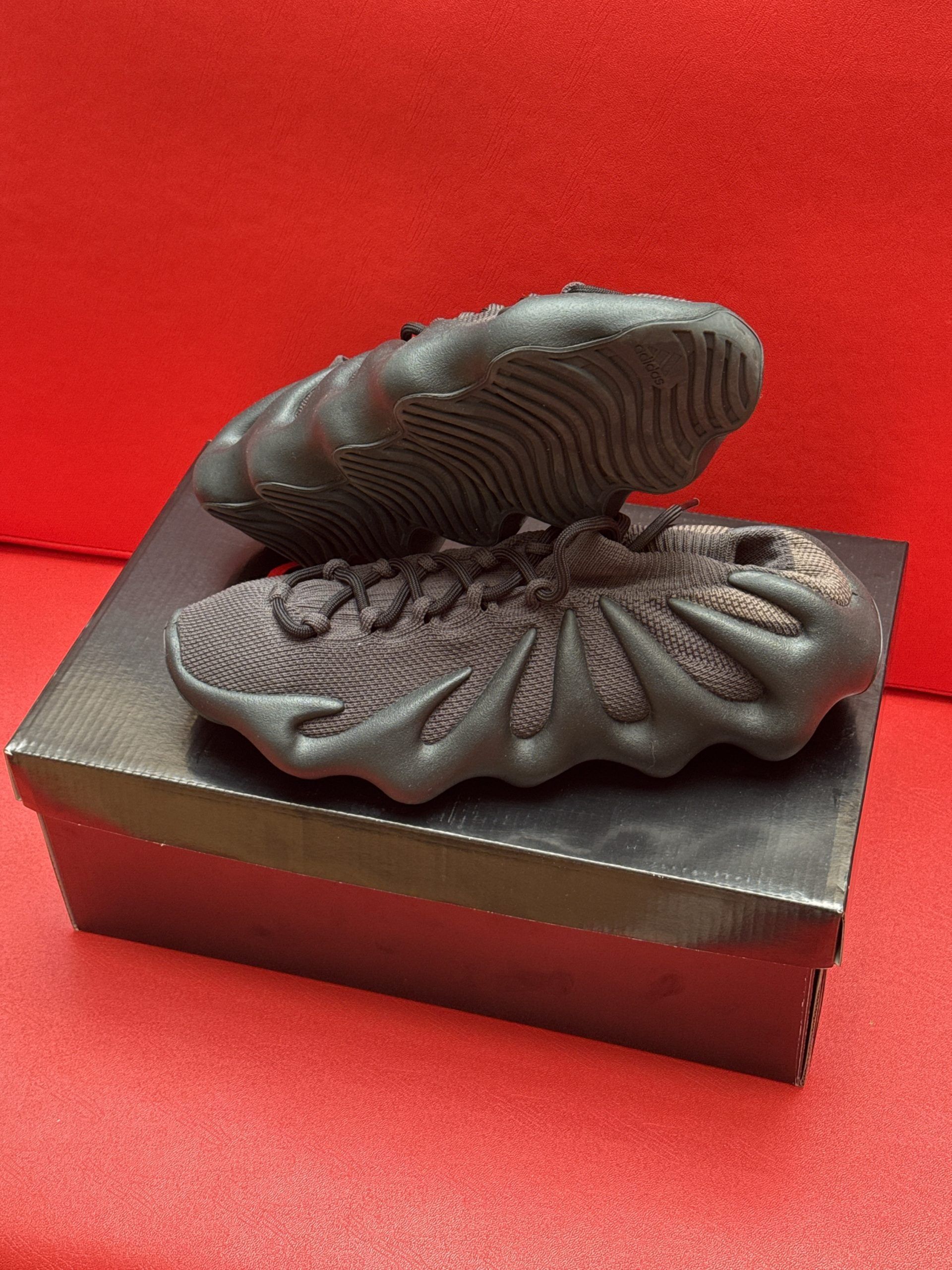 Dark-colored Yeezy 450 sneakers rest on a black shoebox, against a red backdrop.