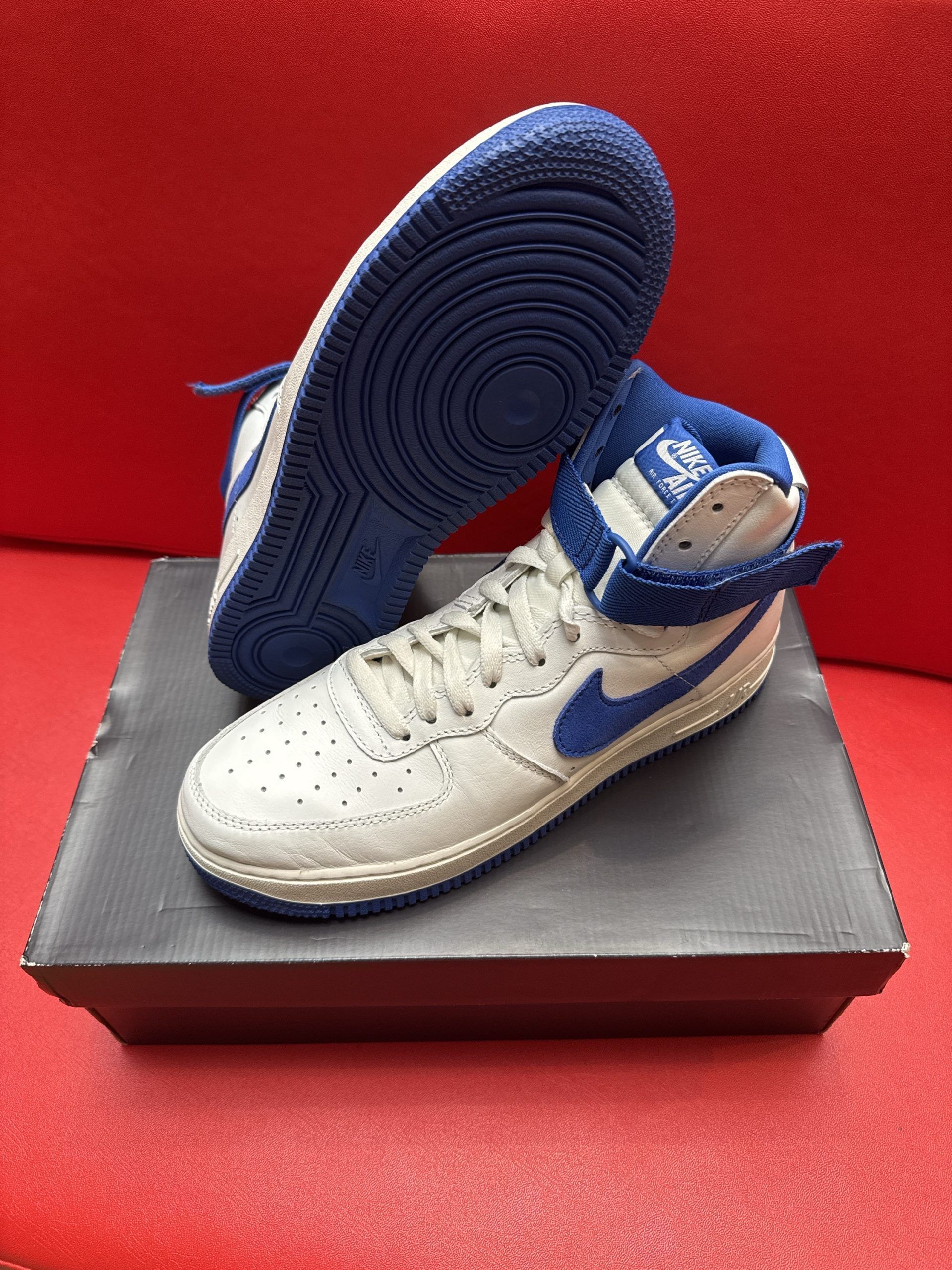 Pair of white and blue Nike high-top sneakers on a black shoebox, set against a red background.