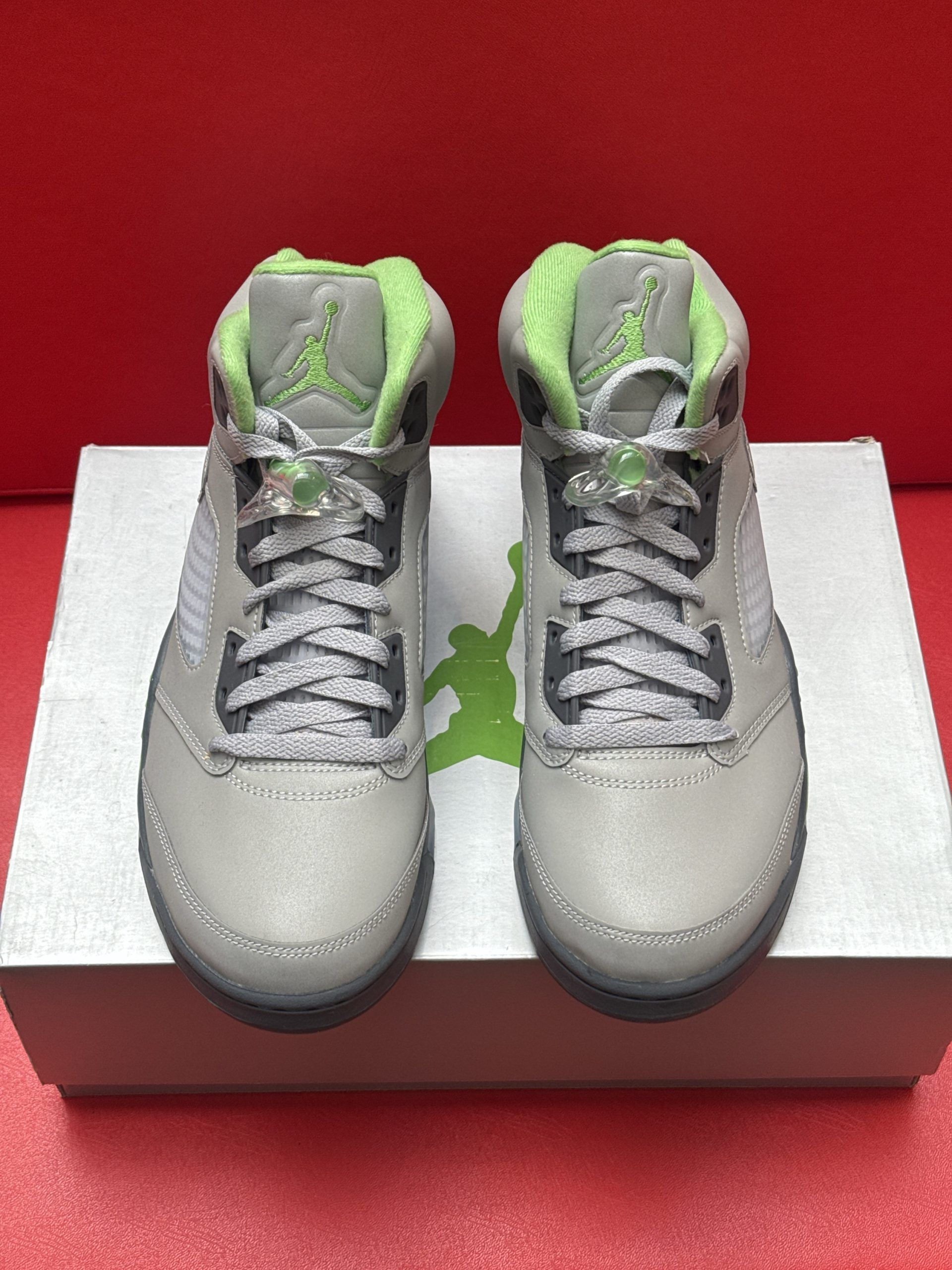 Gray, green, and black Air Jordan 5 sneakers on a white box against a red background.
