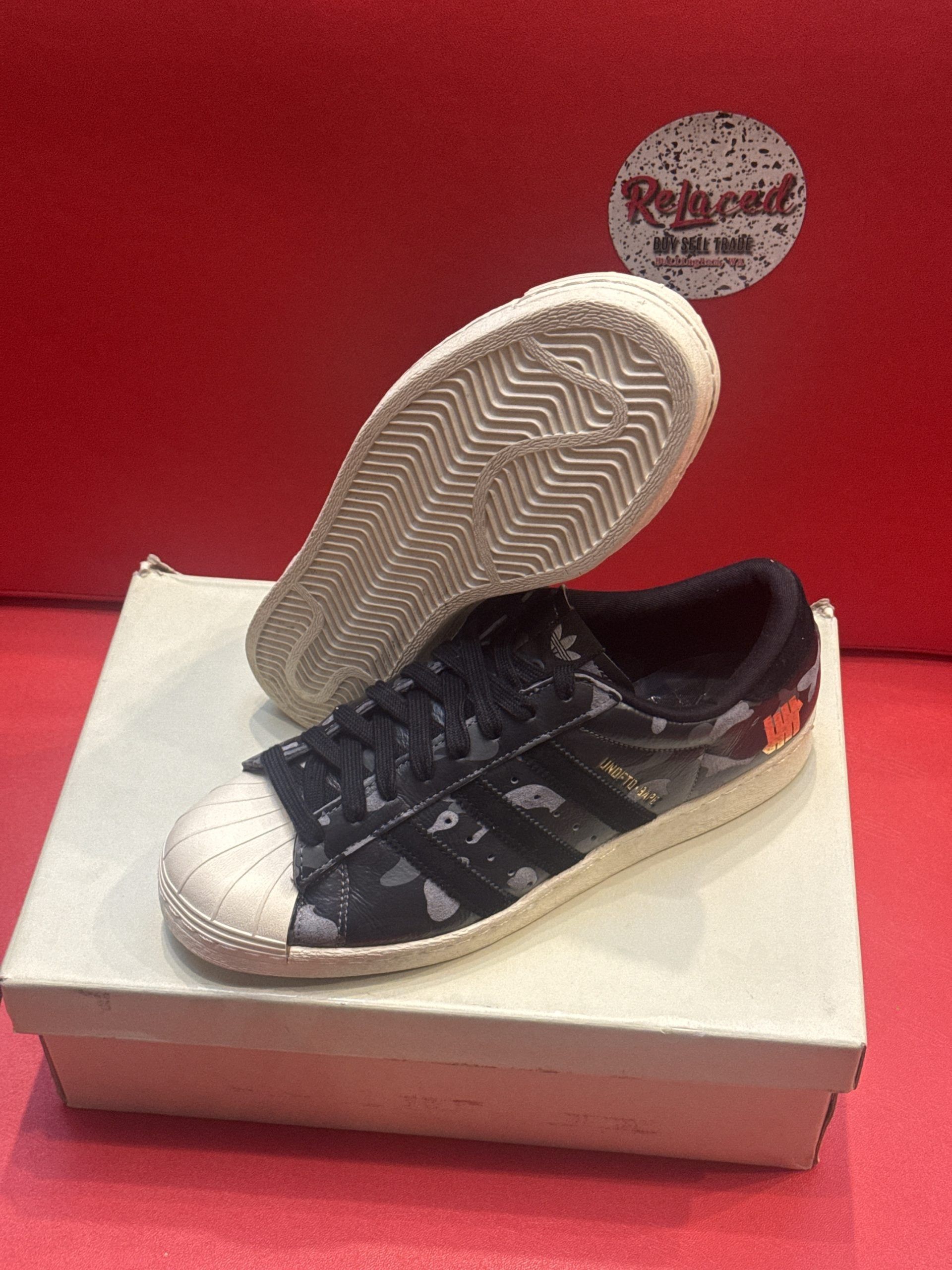 Adidas Superstar sneakers in a box. One sole is up, the other on the box, both white. Black and gray patterns on the shoes.