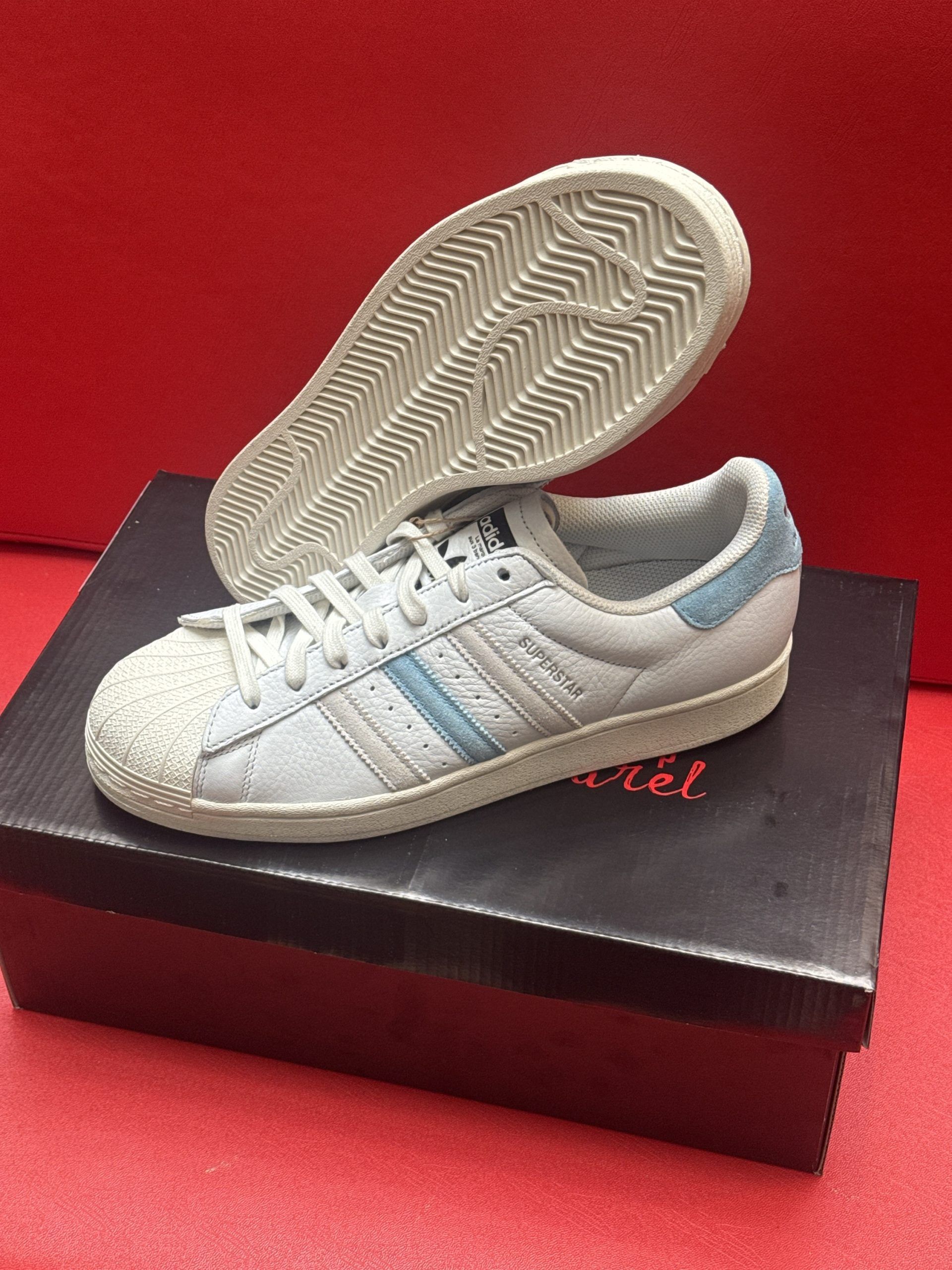 Light blue Adidas Superstar sneakers on a black box, one shoe lifted to show sole, set against a red background.