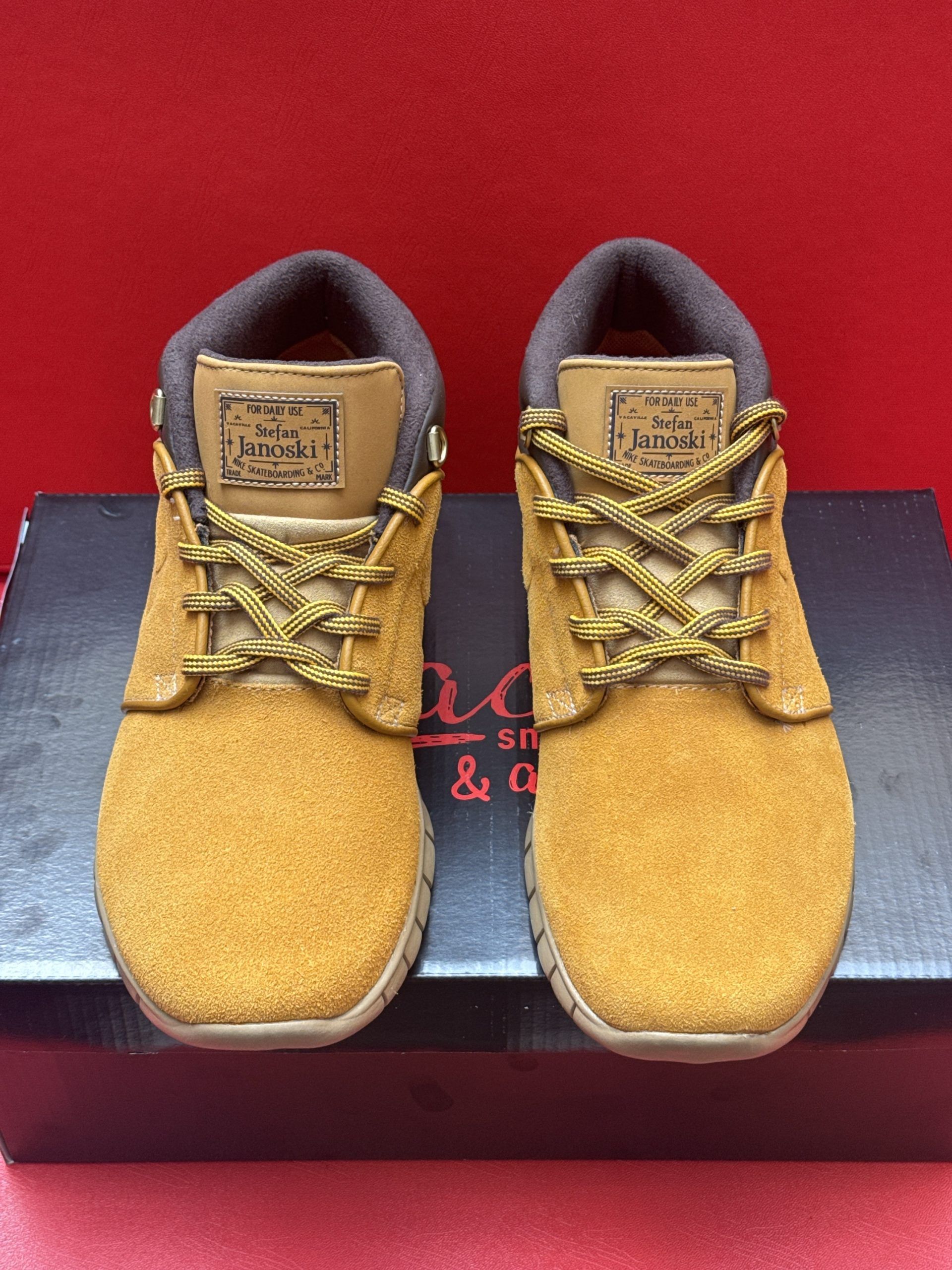 Tan suede Nike shoes with brown trim and laces on a black shoebox, red background.