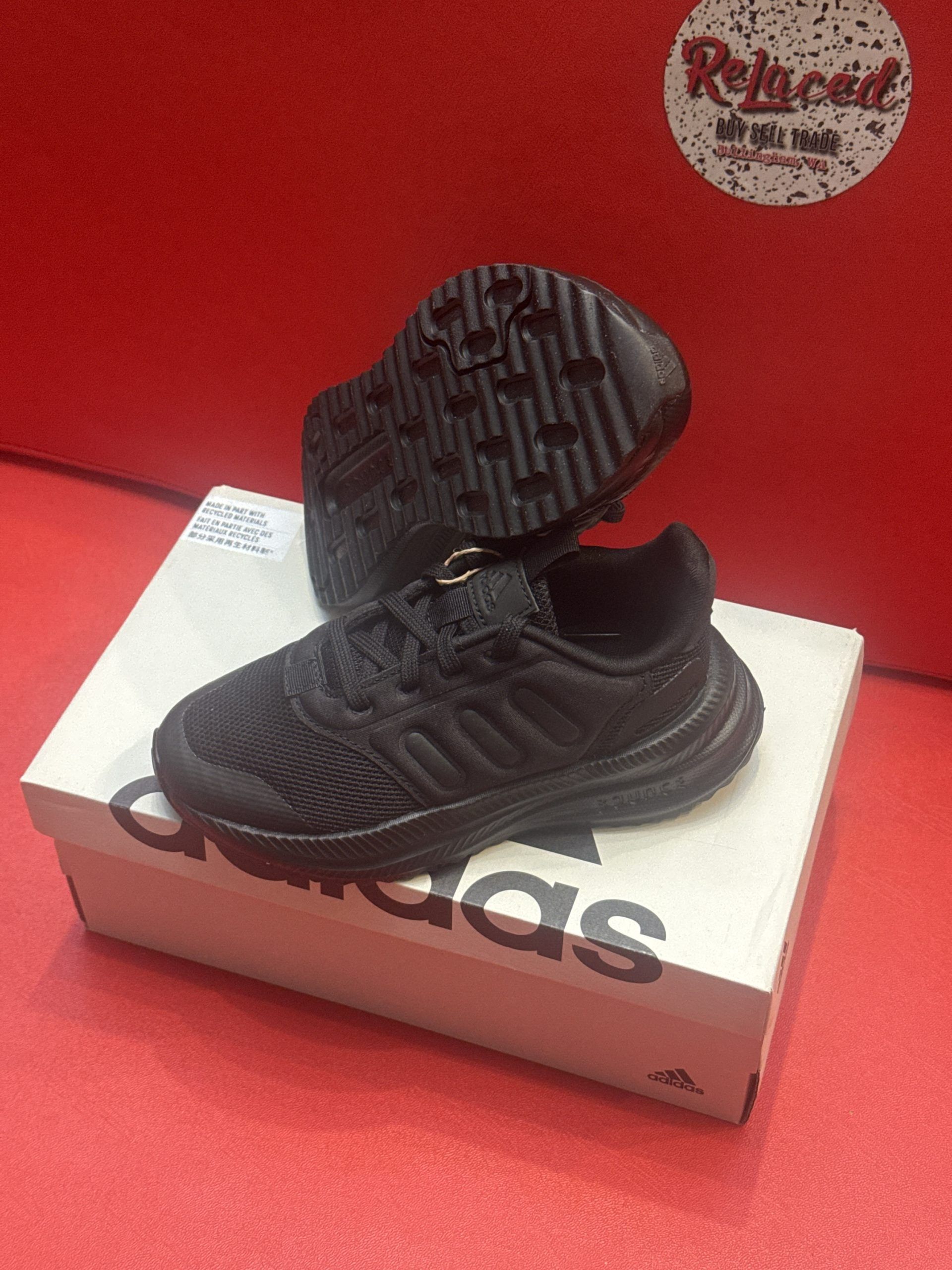 Black Adidas baby shoes on a white Adidas box, set against a red background.