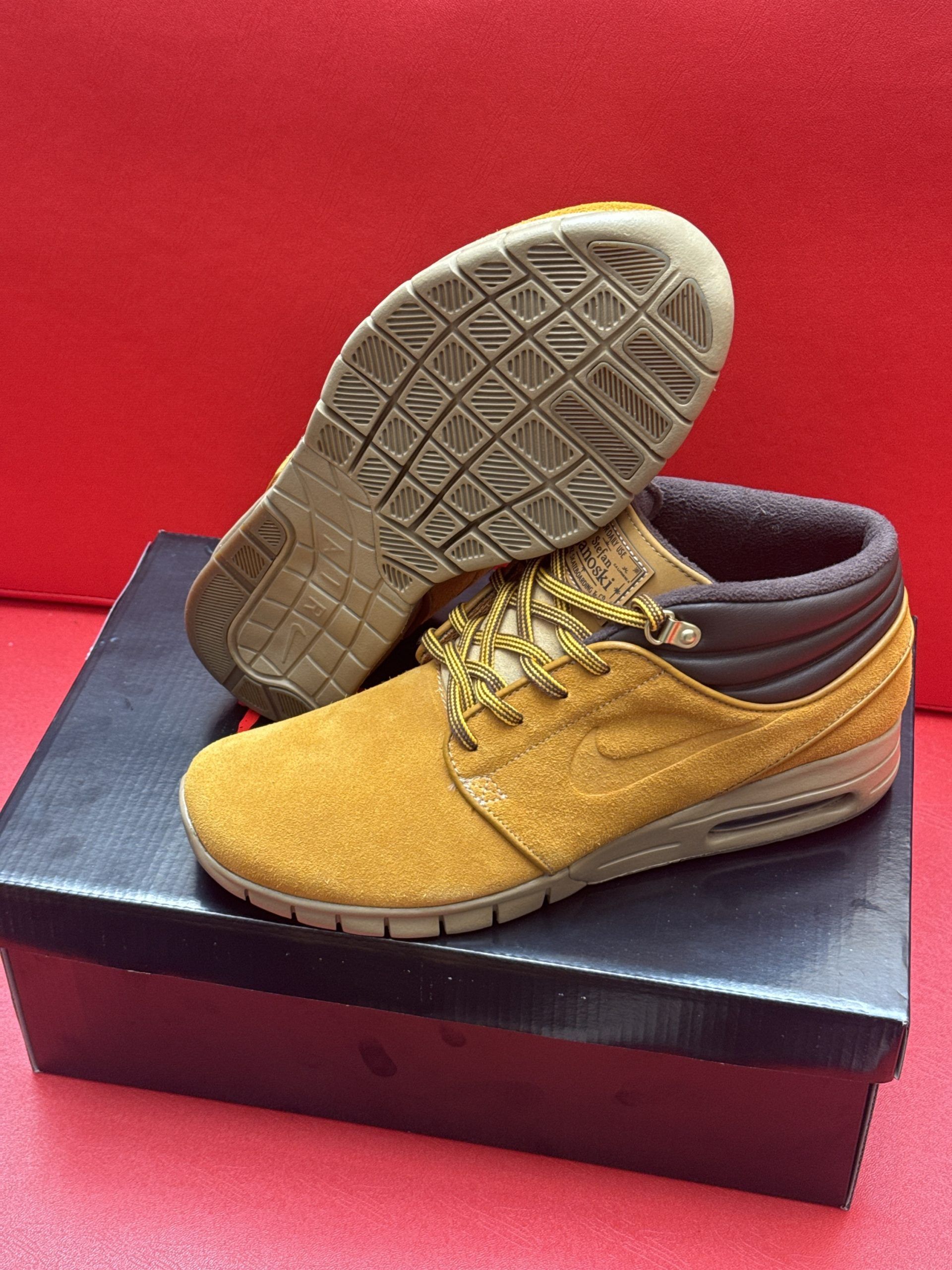 Tan suede Nike shoes with brown trim and laces on a black shoebox, red background.