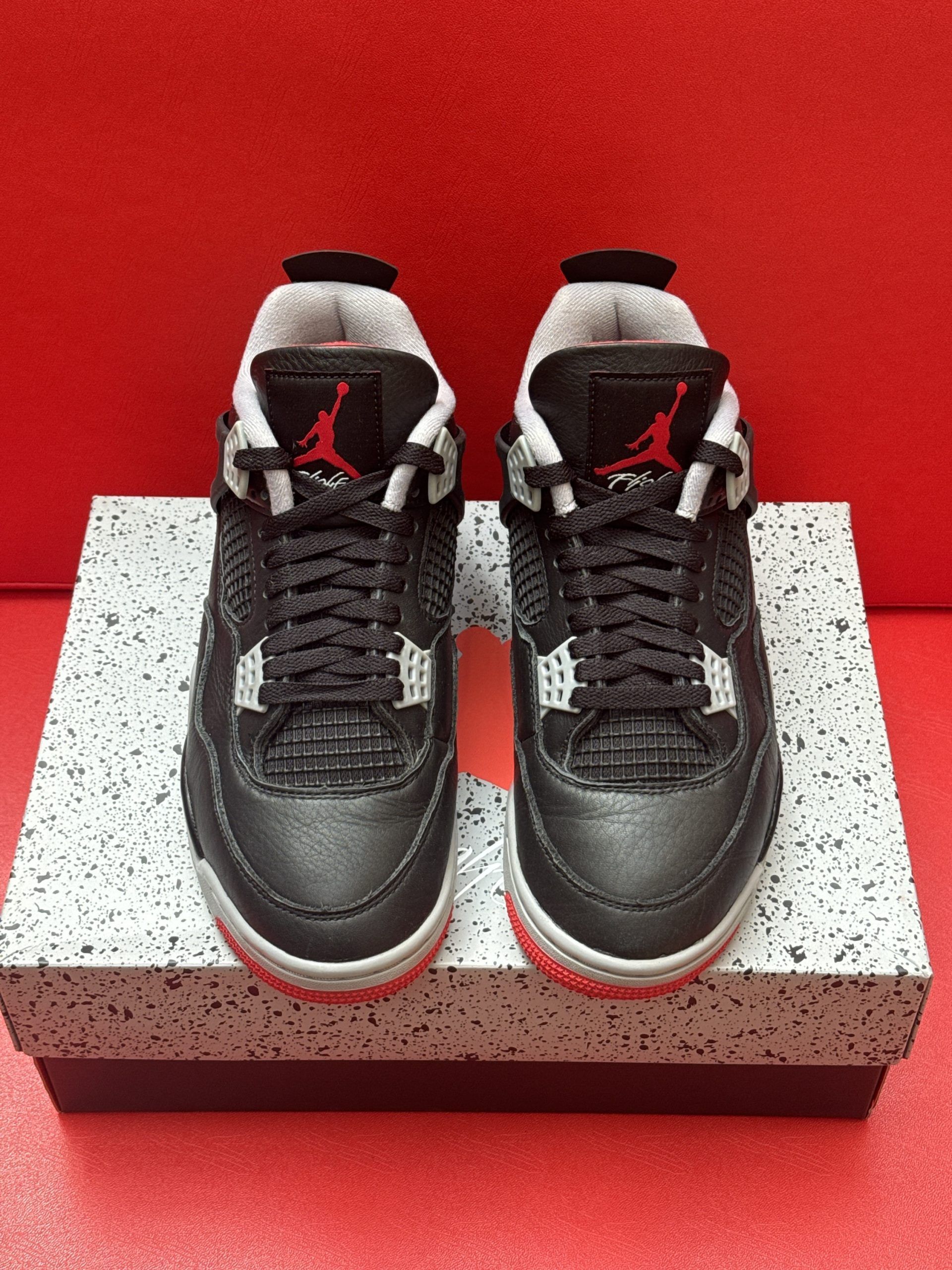 Black, red, and gray Air Jordan 4 infant sneakers on a speckled shoebox. One sneaker's sole is visible.