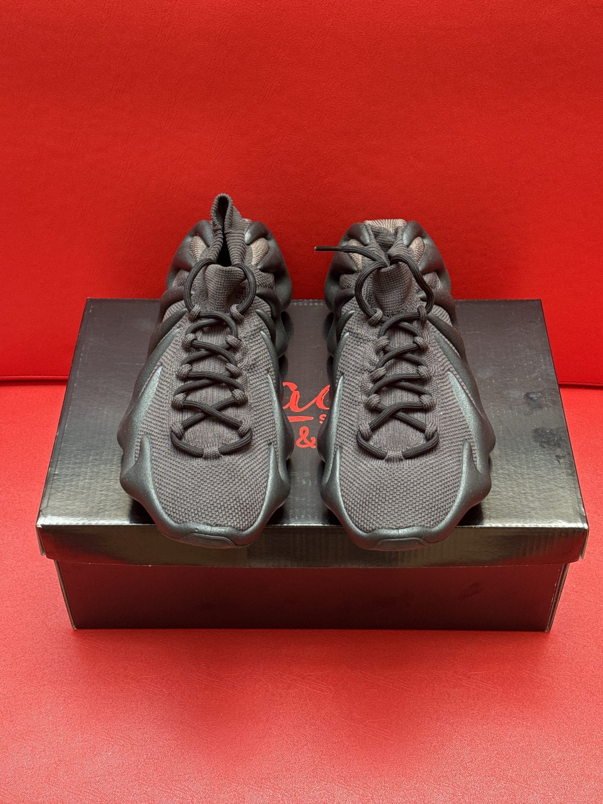 Dark-colored Yeezy 450 sneakers rest on a black shoebox, against a red backdrop.