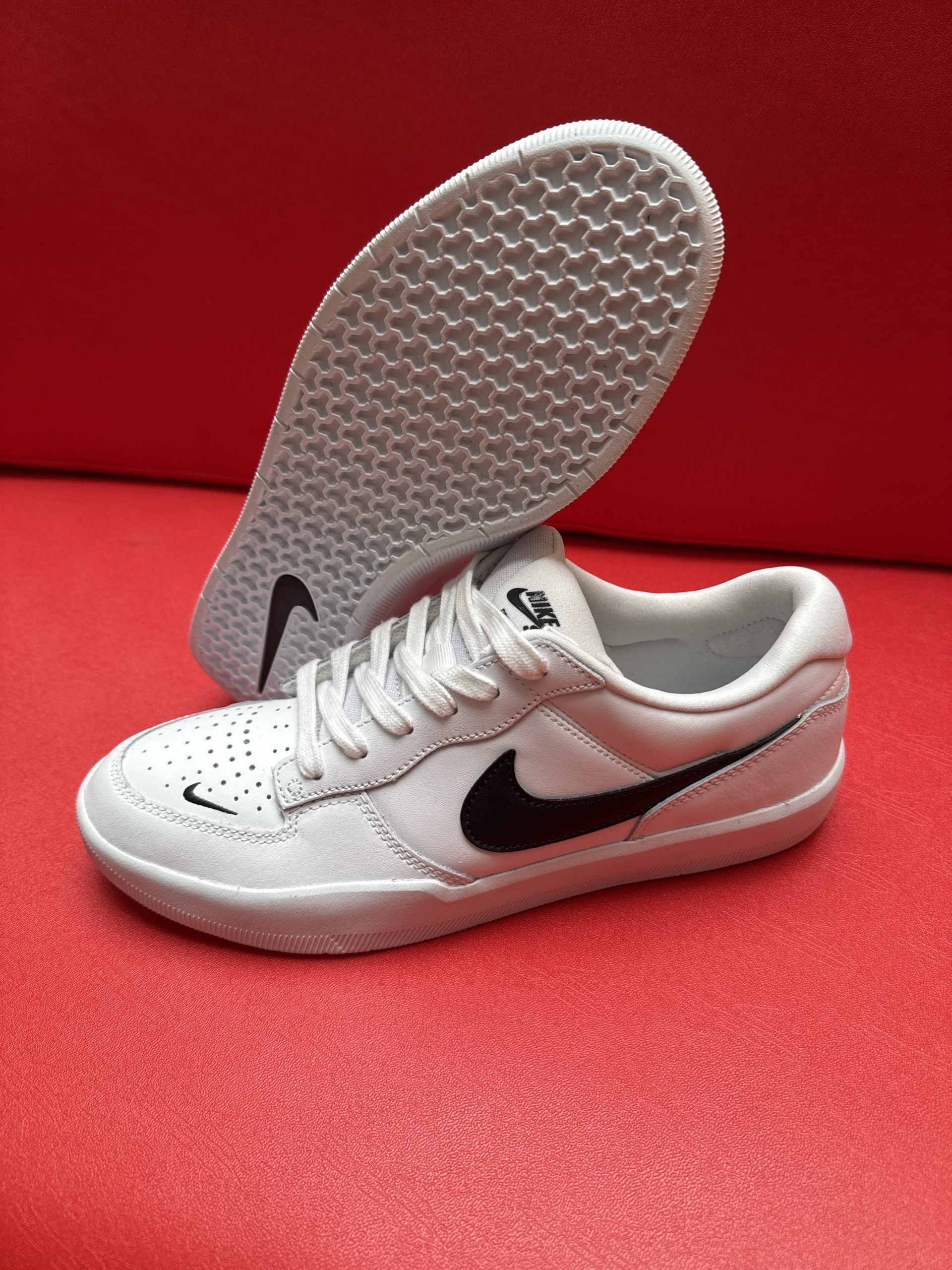 White Nike sneakers with black swoosh on red surface. One shoe is flipped to show the sole.
