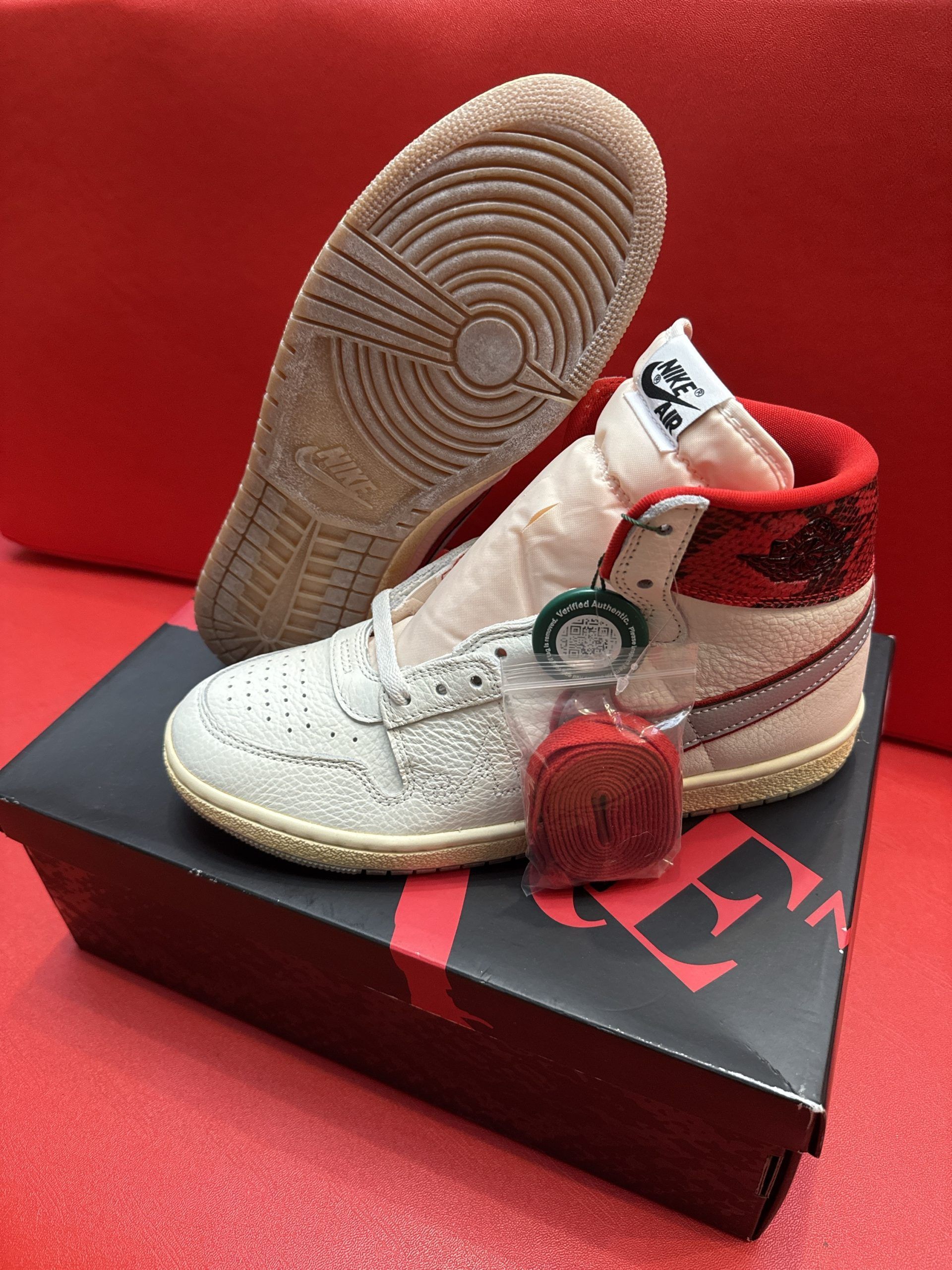 White and red Jordan 1 sneaker with shoebox, sole displayed, on red surface.