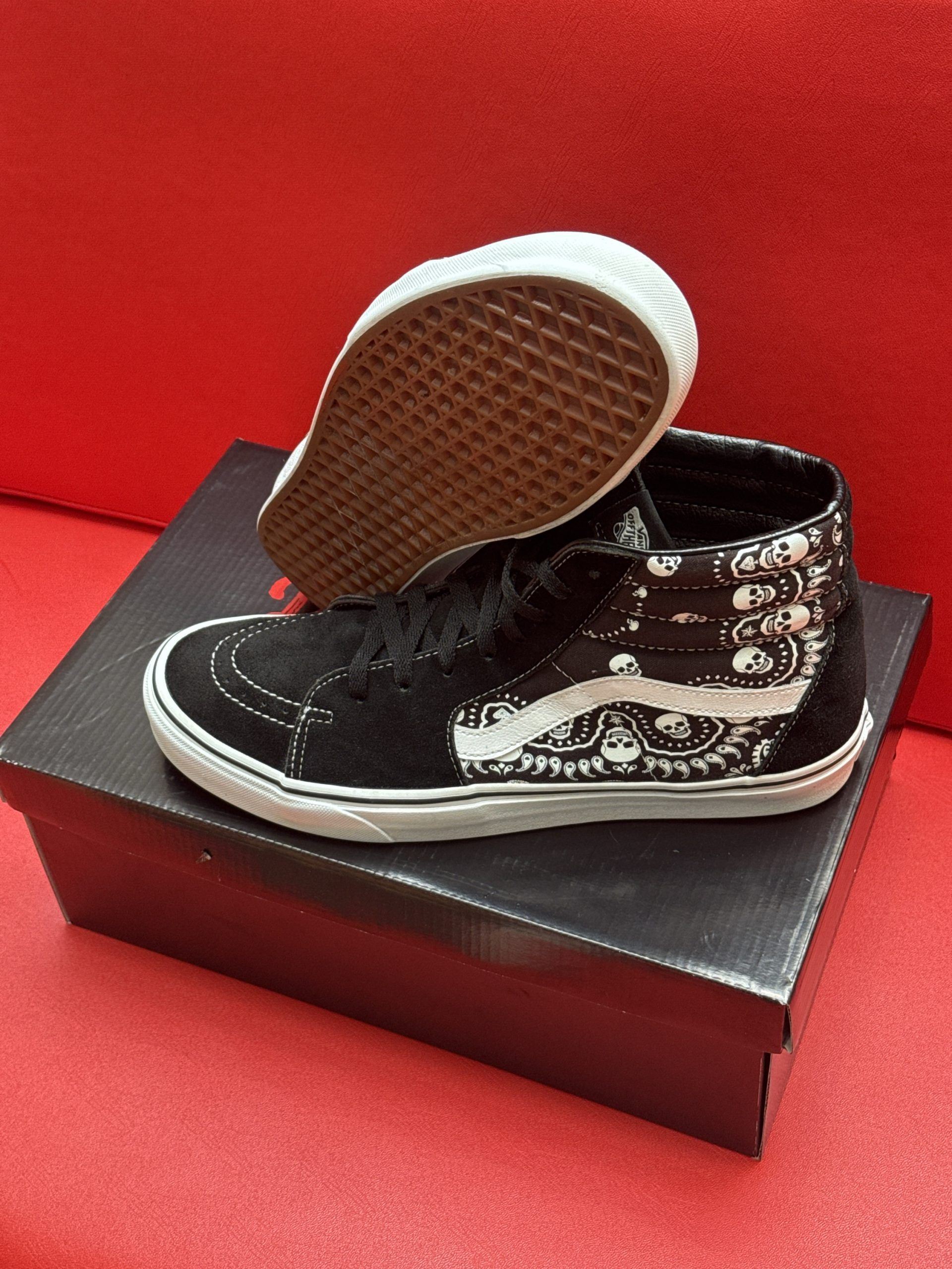 Black and white Vans SK8-Hi sneakers with a paisley pattern sit on a black shoebox against a red background.