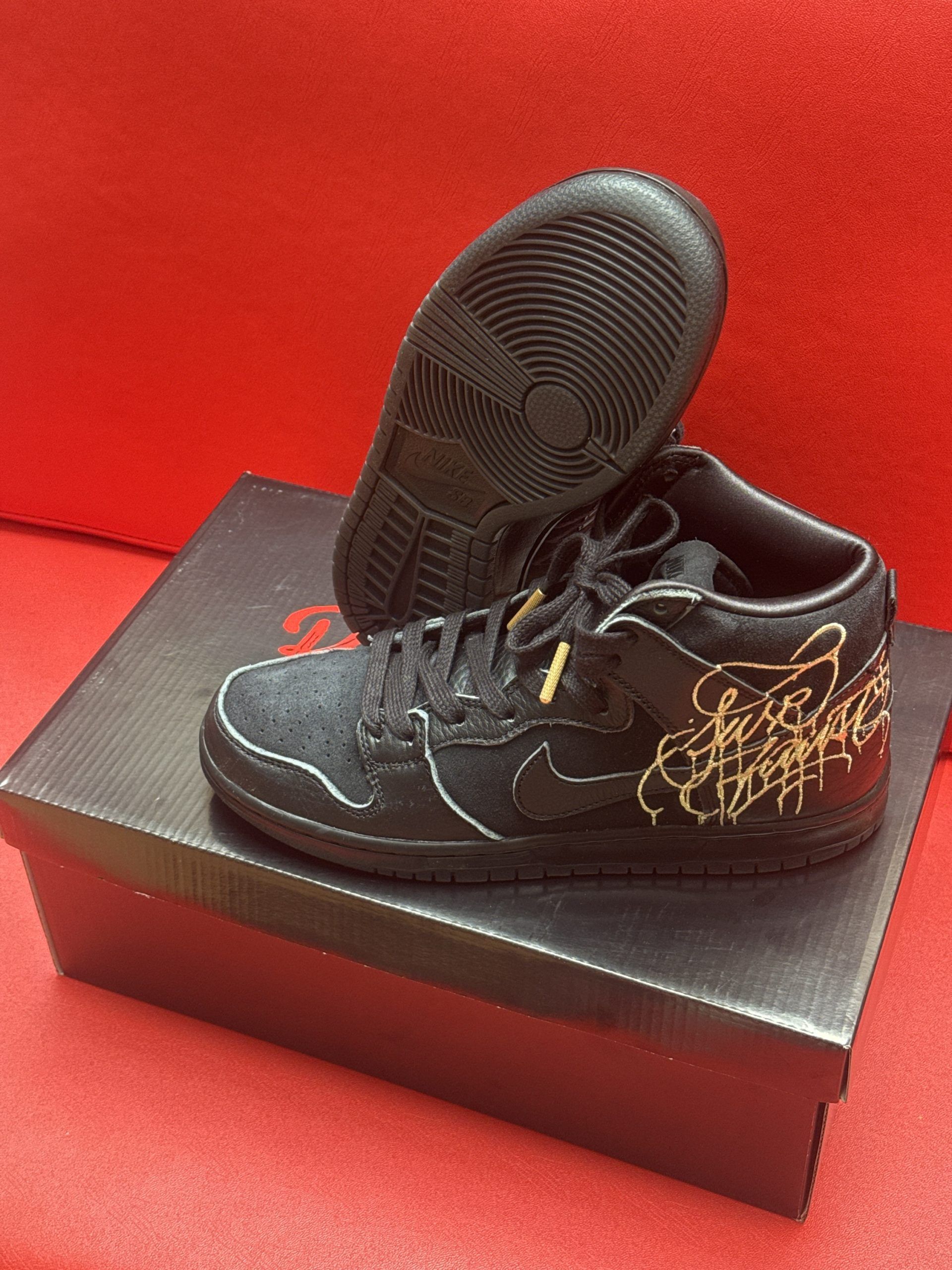 Black Nike SB Dunk High sneakers with gold accents, on a black shoebox.