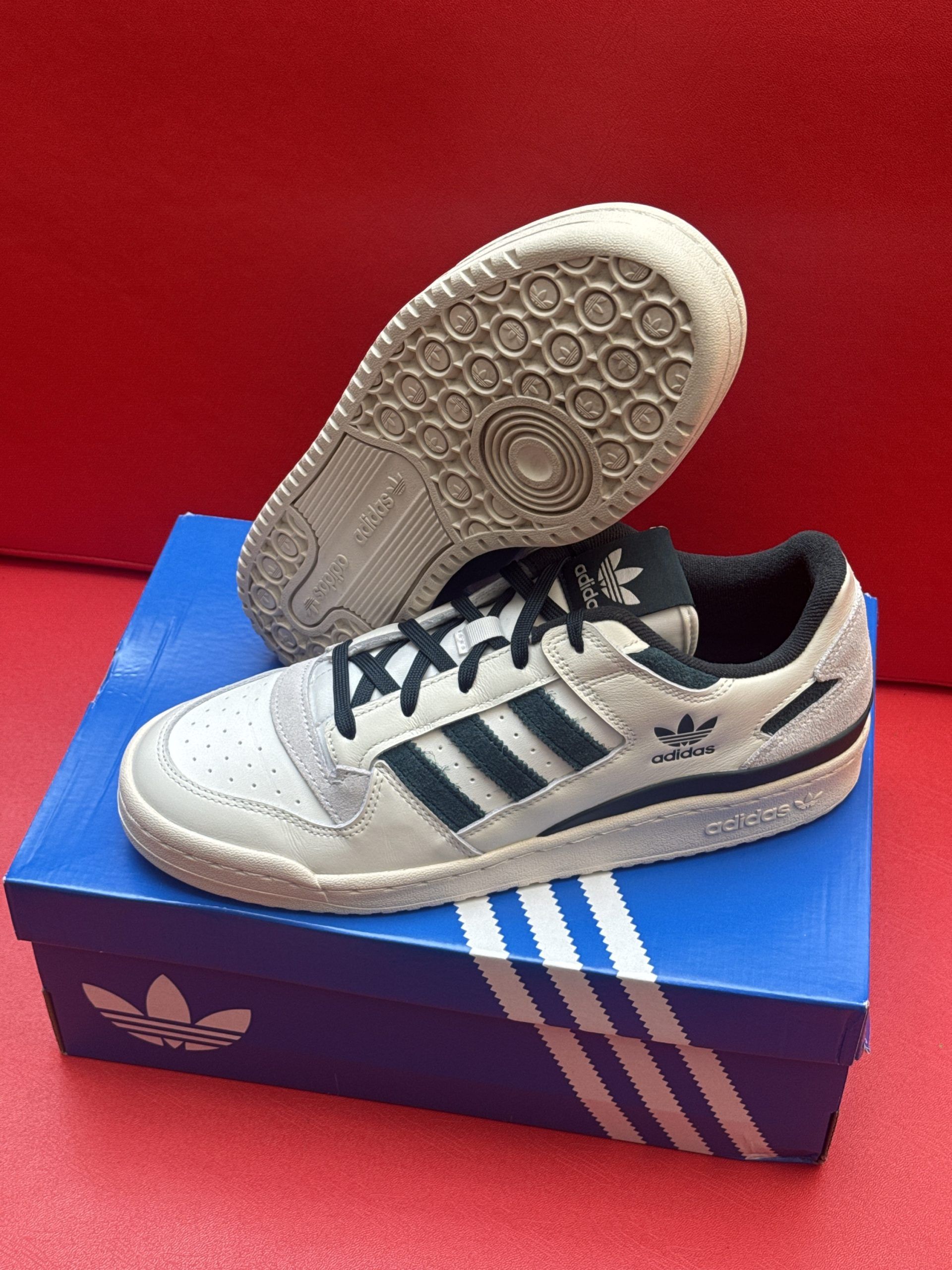 Adidas Forum low sneakers in off-white with dark green stripes on the box against a red background.