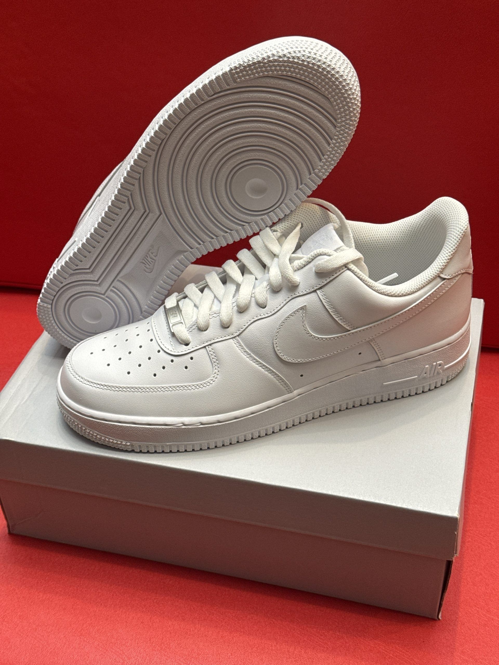 White Nike Air Force 1 sneakers on a white box, one shoe upside down showing the sole; red background.