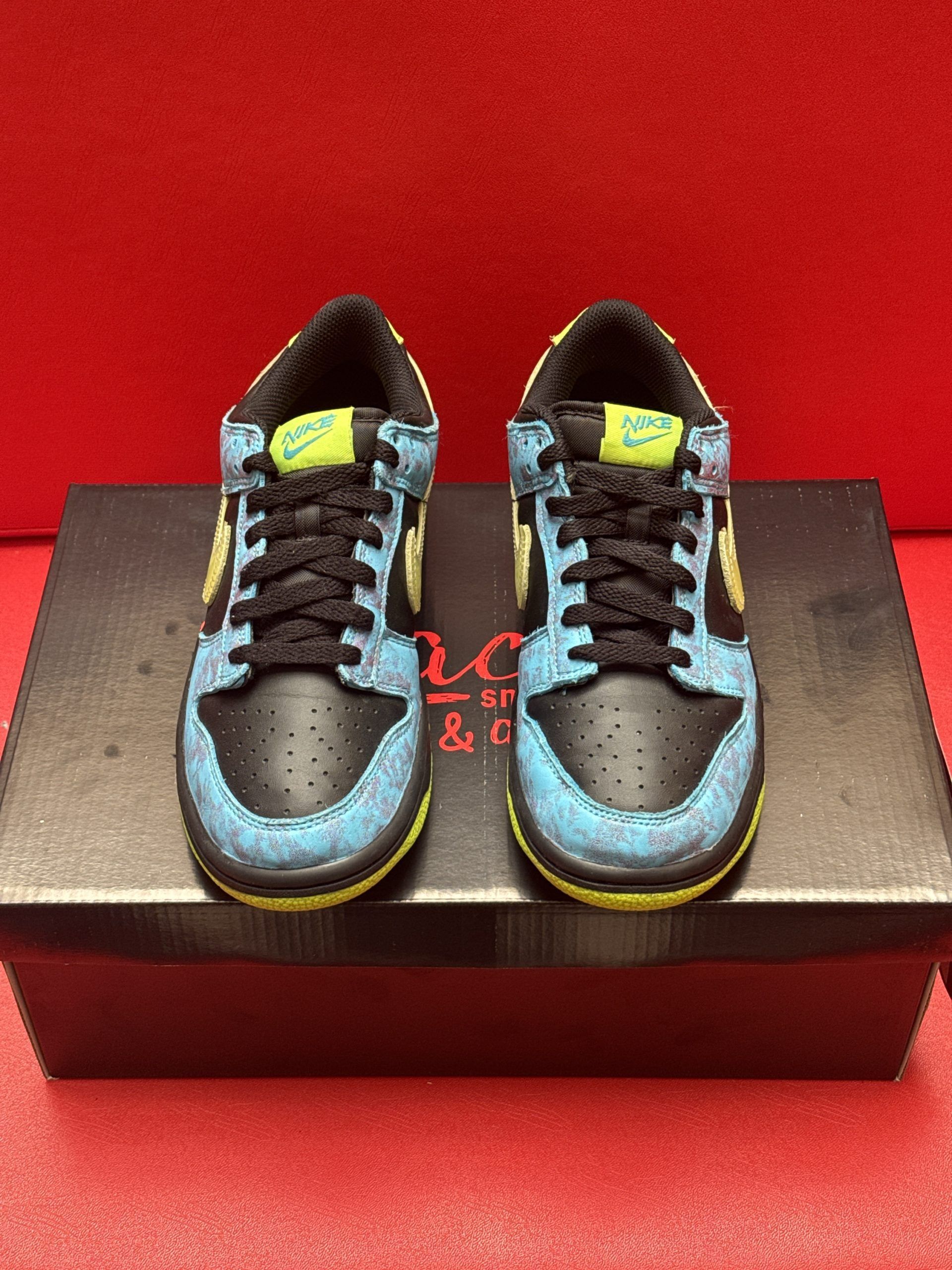 Nike Dunk low sneaker in black, blue, and yellow on a black shoe box with a red background.
