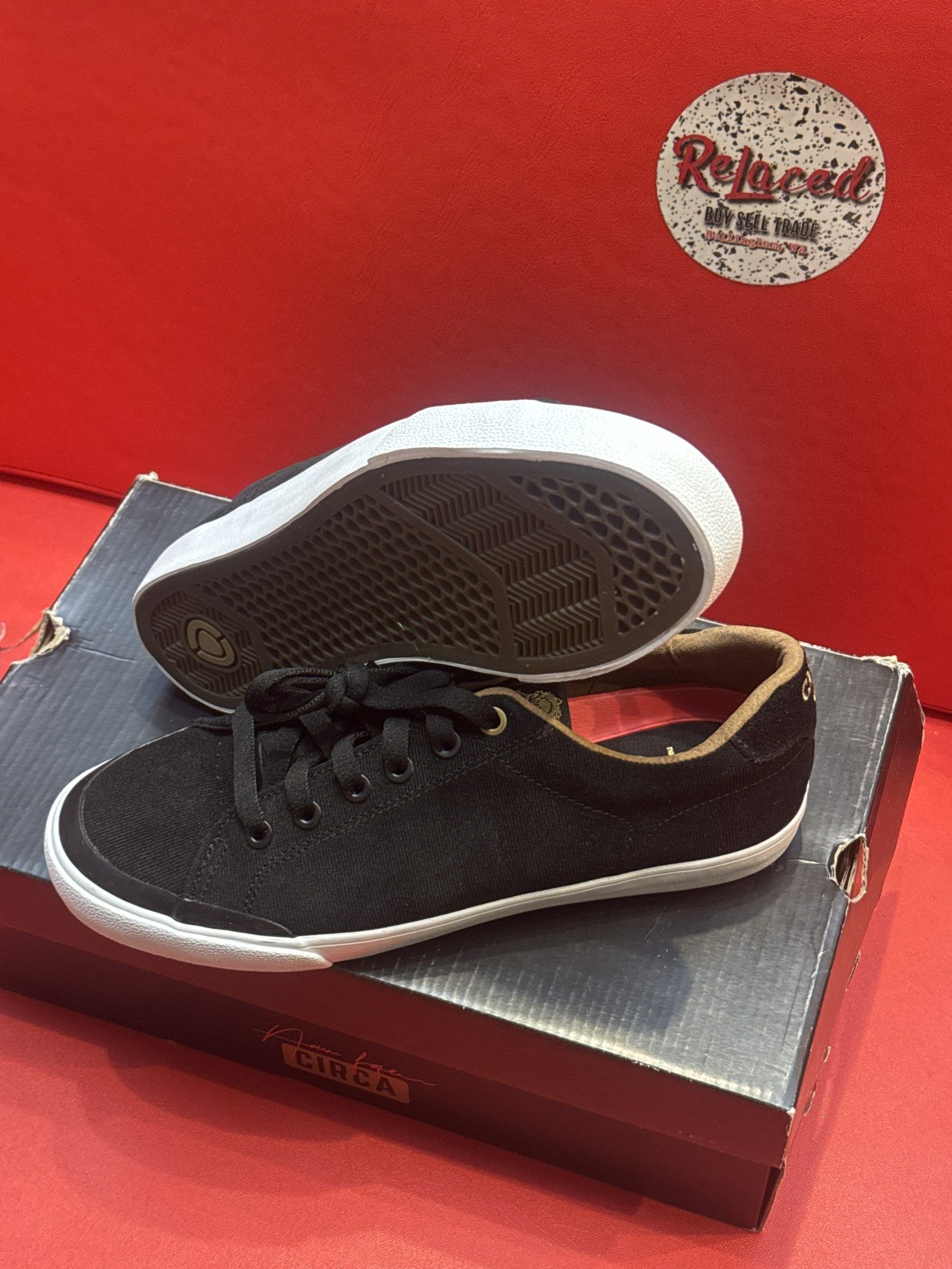 Black sneakers with white soles, laces, and a brown interior, atop a shoebox on a red surface.
