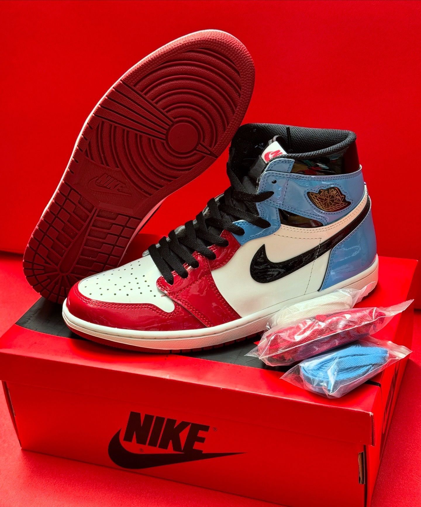 Red, white, and blue Air Jordan 1 sneaker on a red box with extra laces and a red sole against a red backdrop.