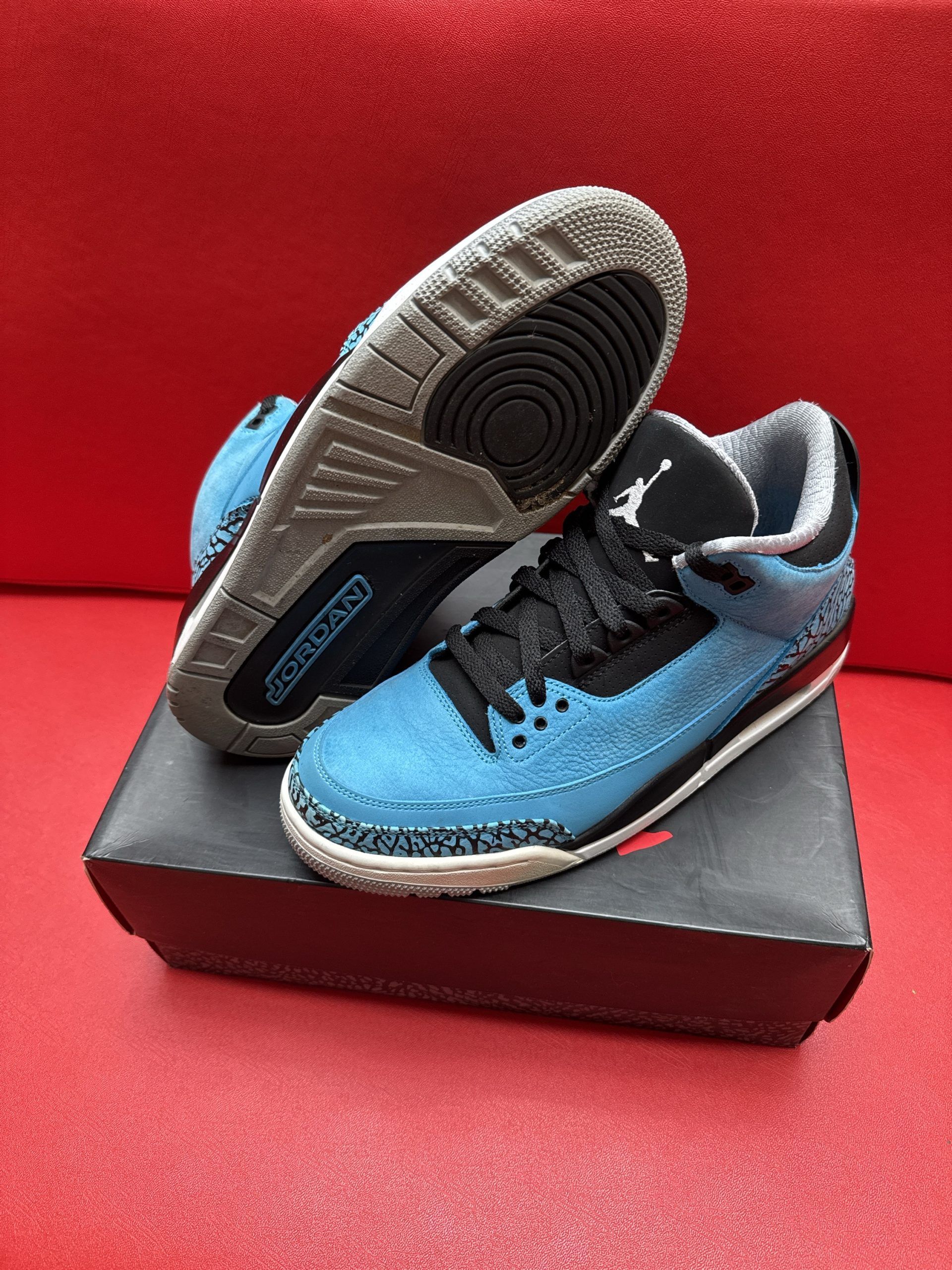 Baby blue and black Air Jordan 3 sneakers on a black box, gray sole. Red background.
