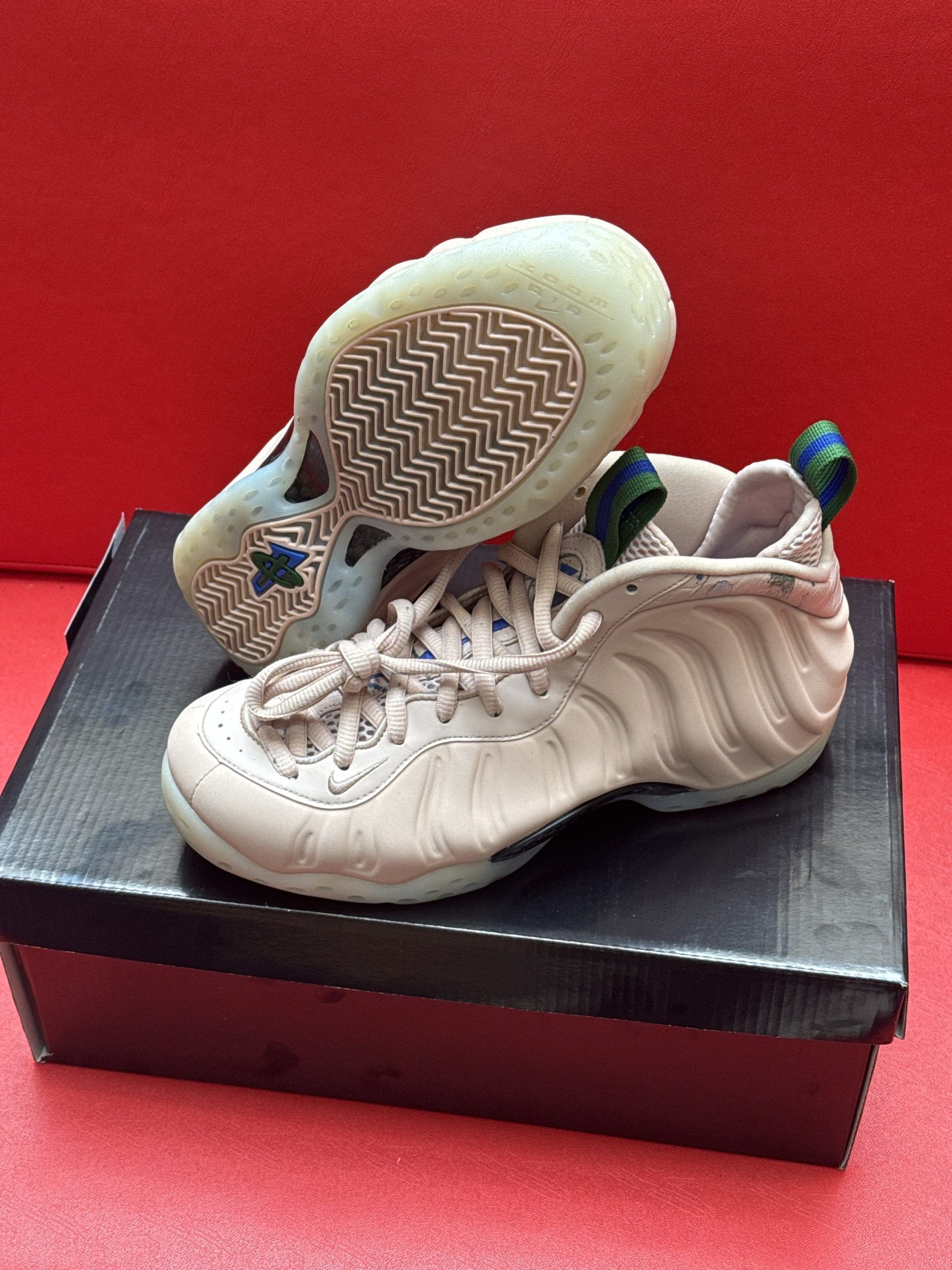 Tan Nike Foamposite sneakers on a black shoebox, sole up.  Green and blue details, red background.
