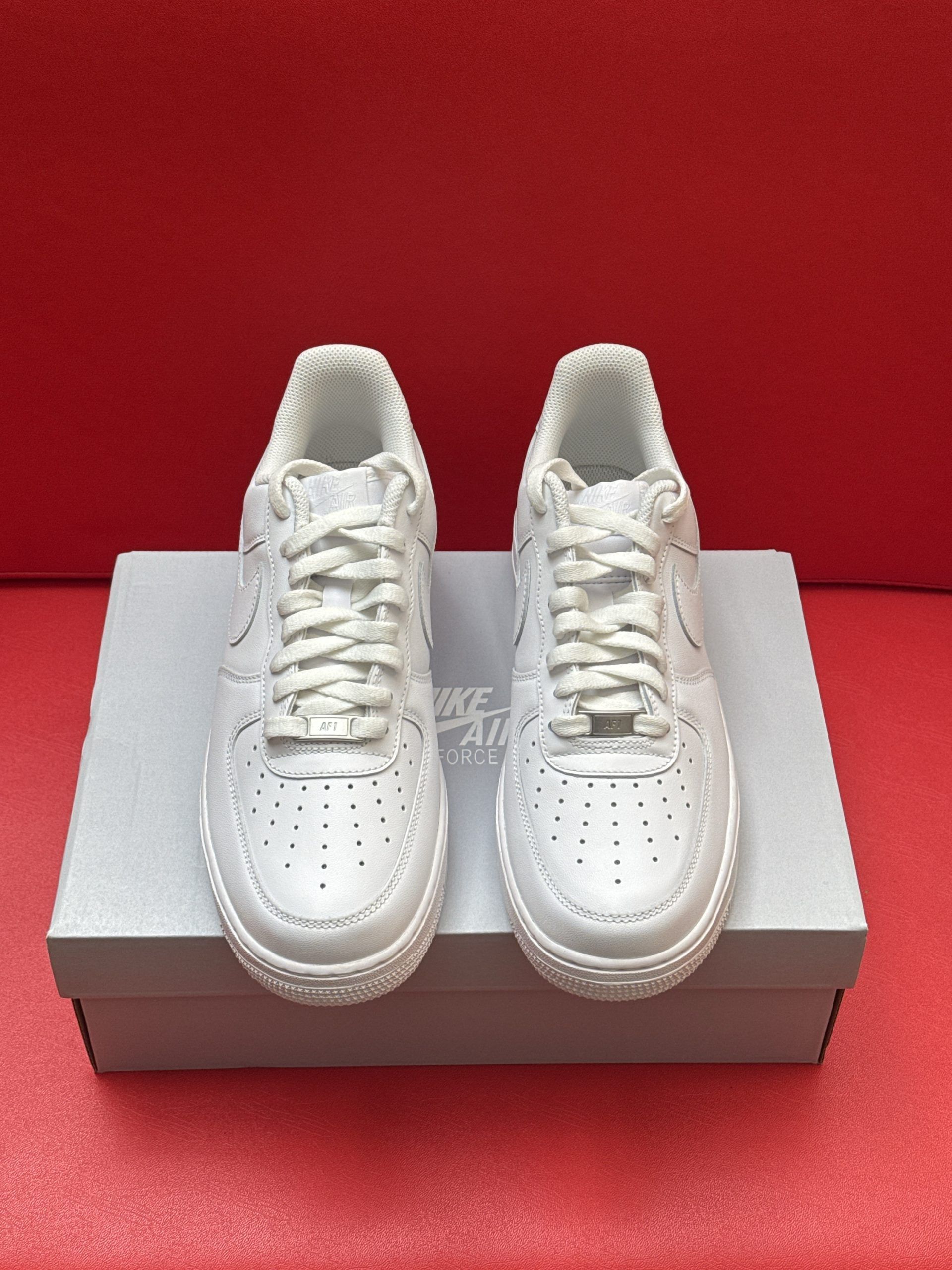 White Nike Air Force 1 sneakers on top of a shoe box, sole facing upwards. Red background.