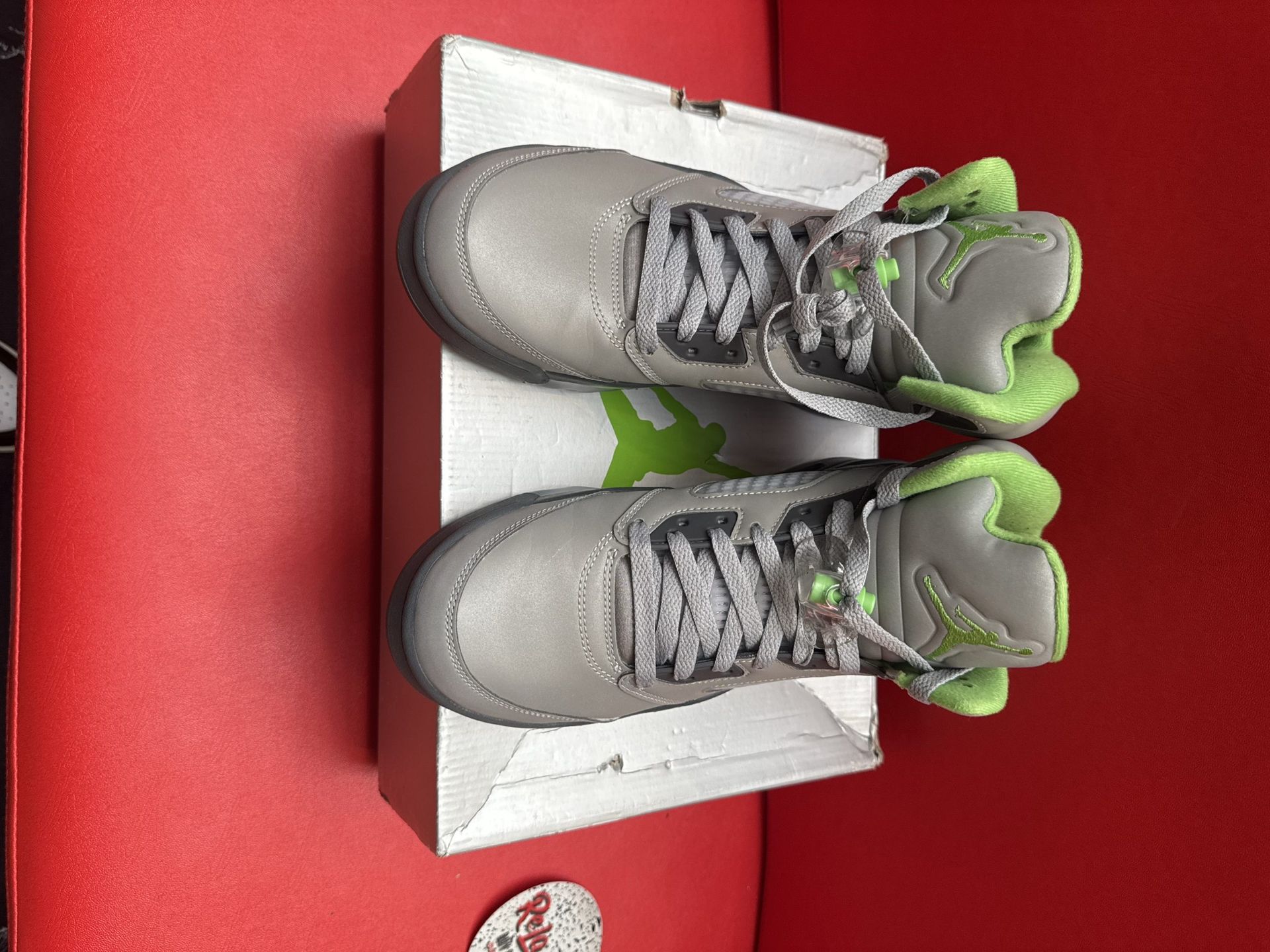 Gray and green Air Jordan 5 sneakers on a white box, sole visible, set on a red surface.