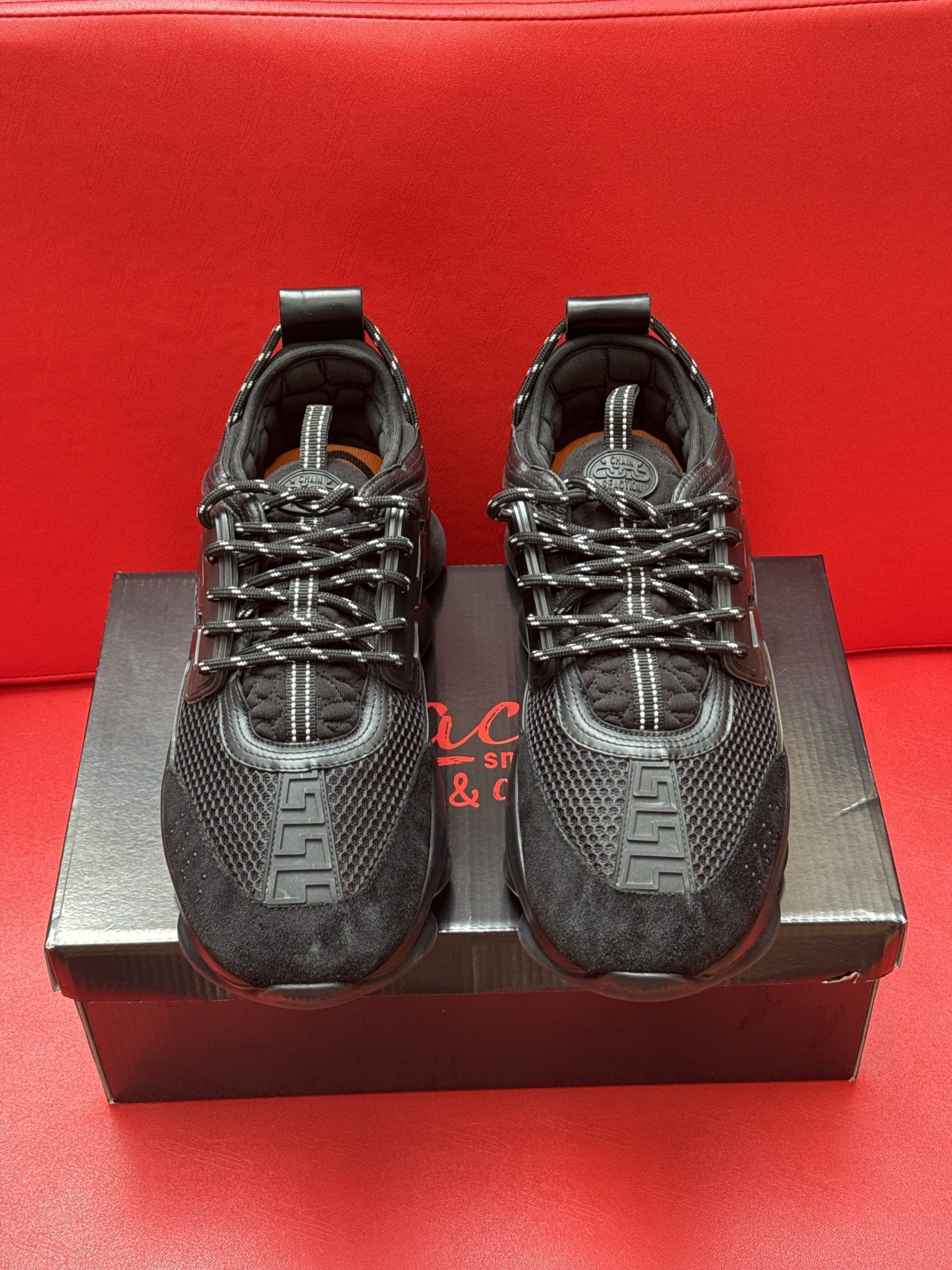 Black Versace Chain Reaction sneakers on a black box, with a red background.