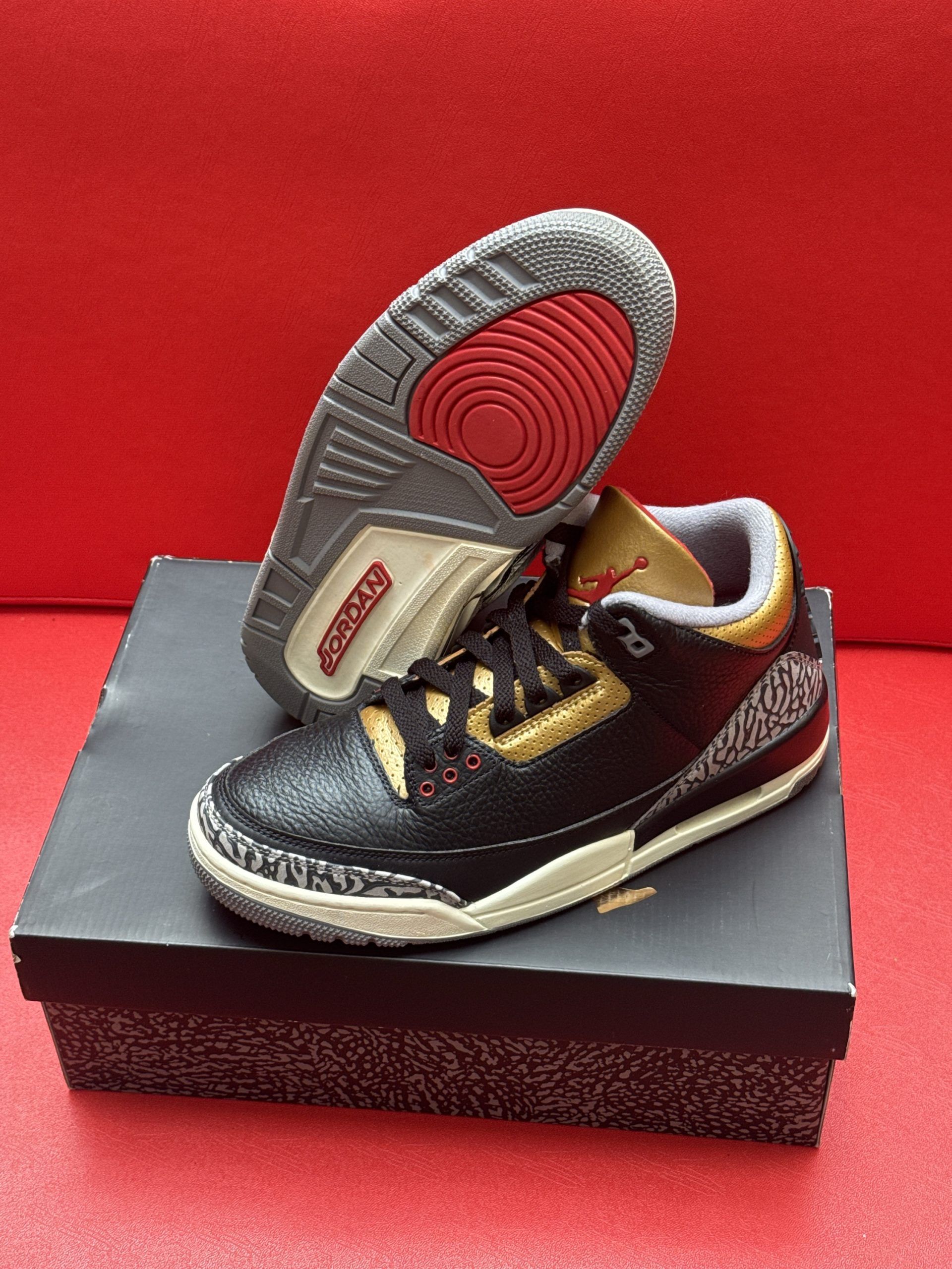 Black, gold, and red Air Jordan 3 sneakers on a black box, sole visible, set against a red background.