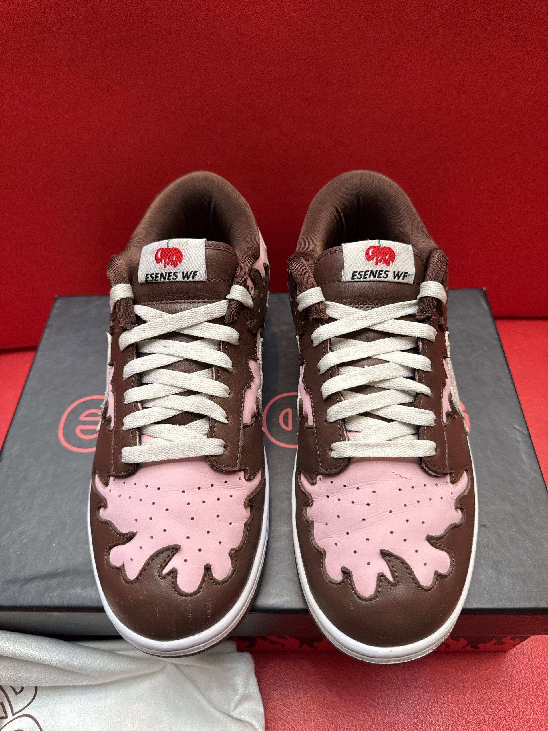 Brown and pink sneakers with flame design, sitting on a black box, and white bag.