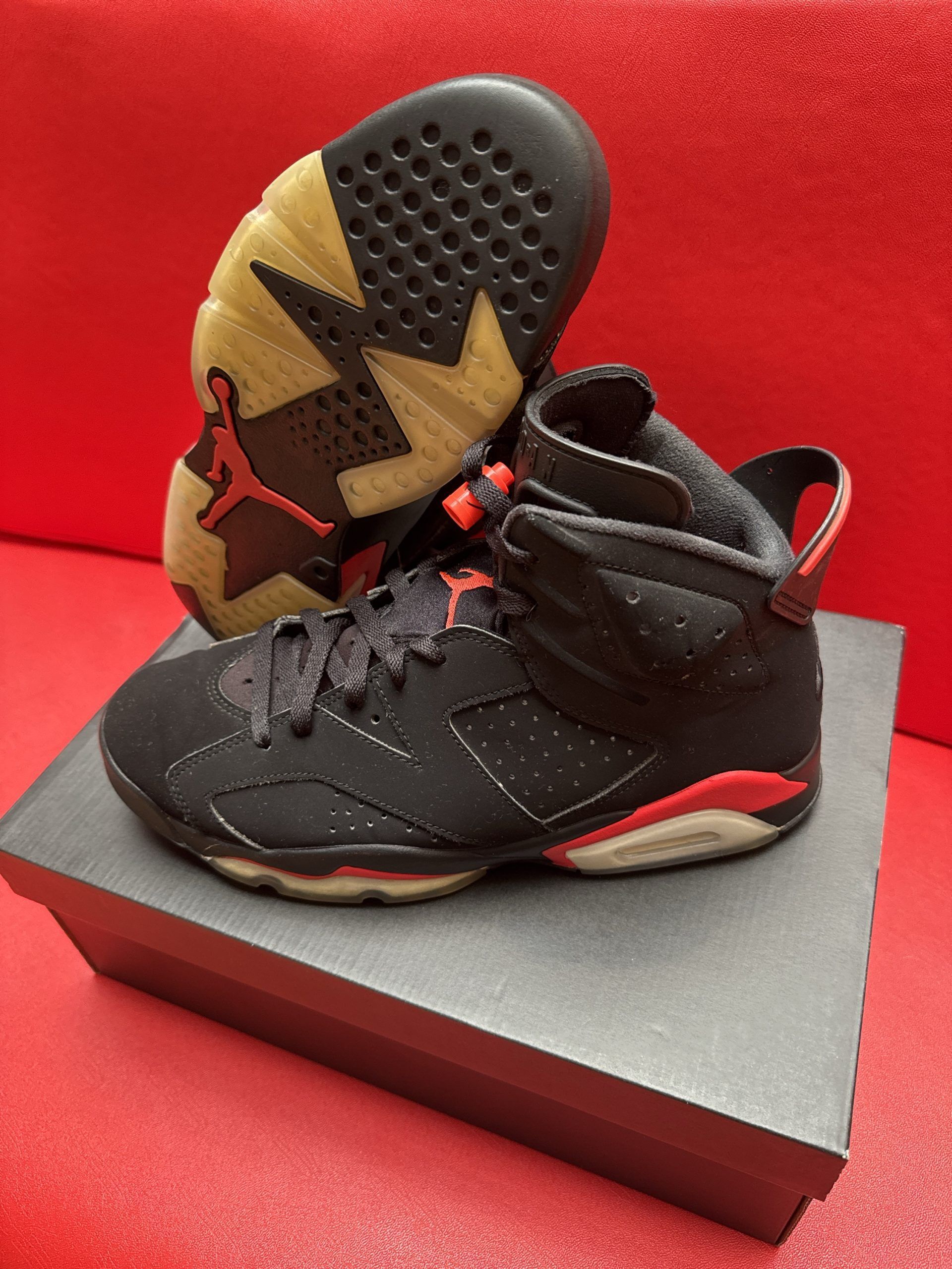 Black and red Air Jordan 6 sneakers on a box, with one turned to show the sole on a red surface.